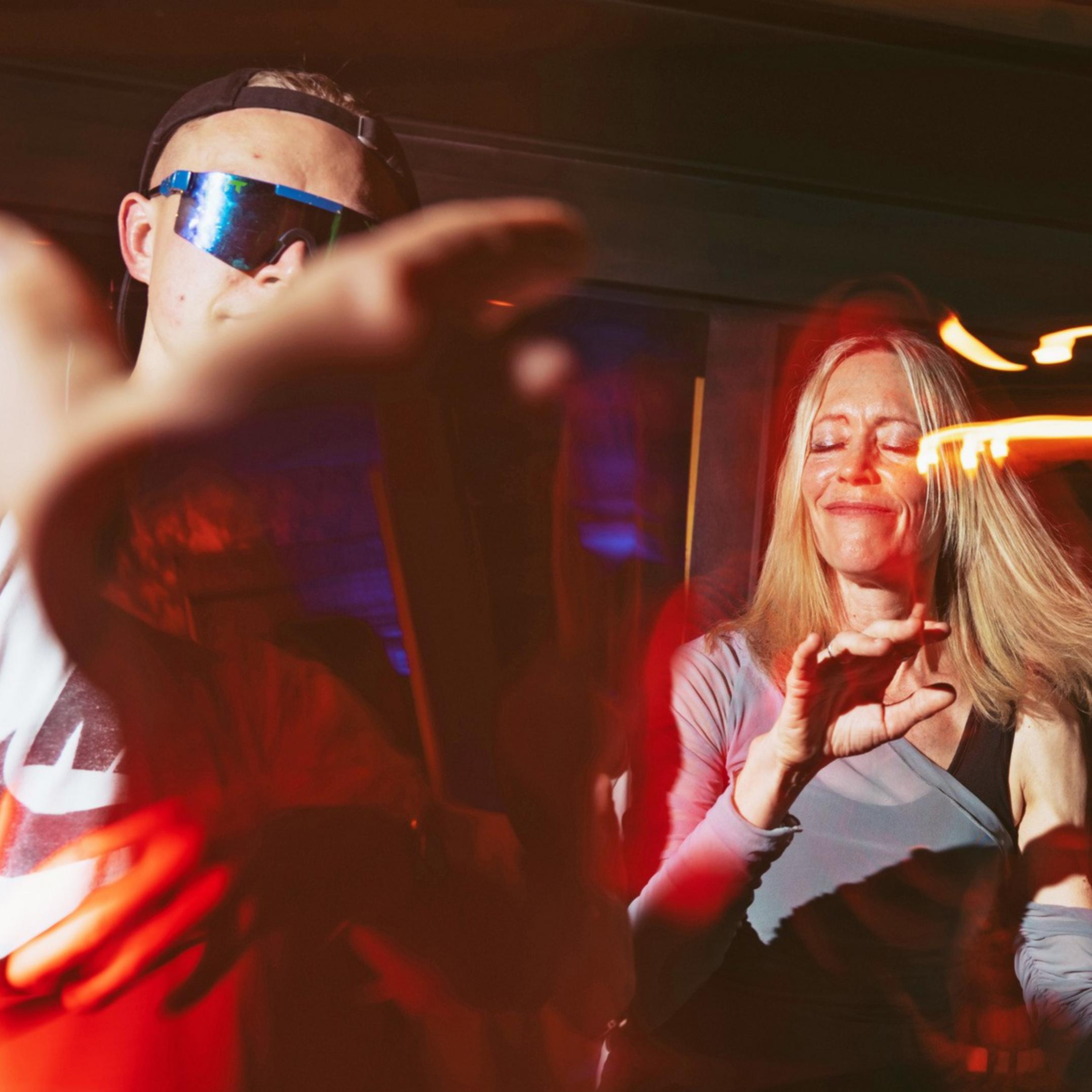 People are dancing energetically in a dimly lit space with colorful lights. A woman in the center smiles with her eyes closed, surrounded by blurred motion.