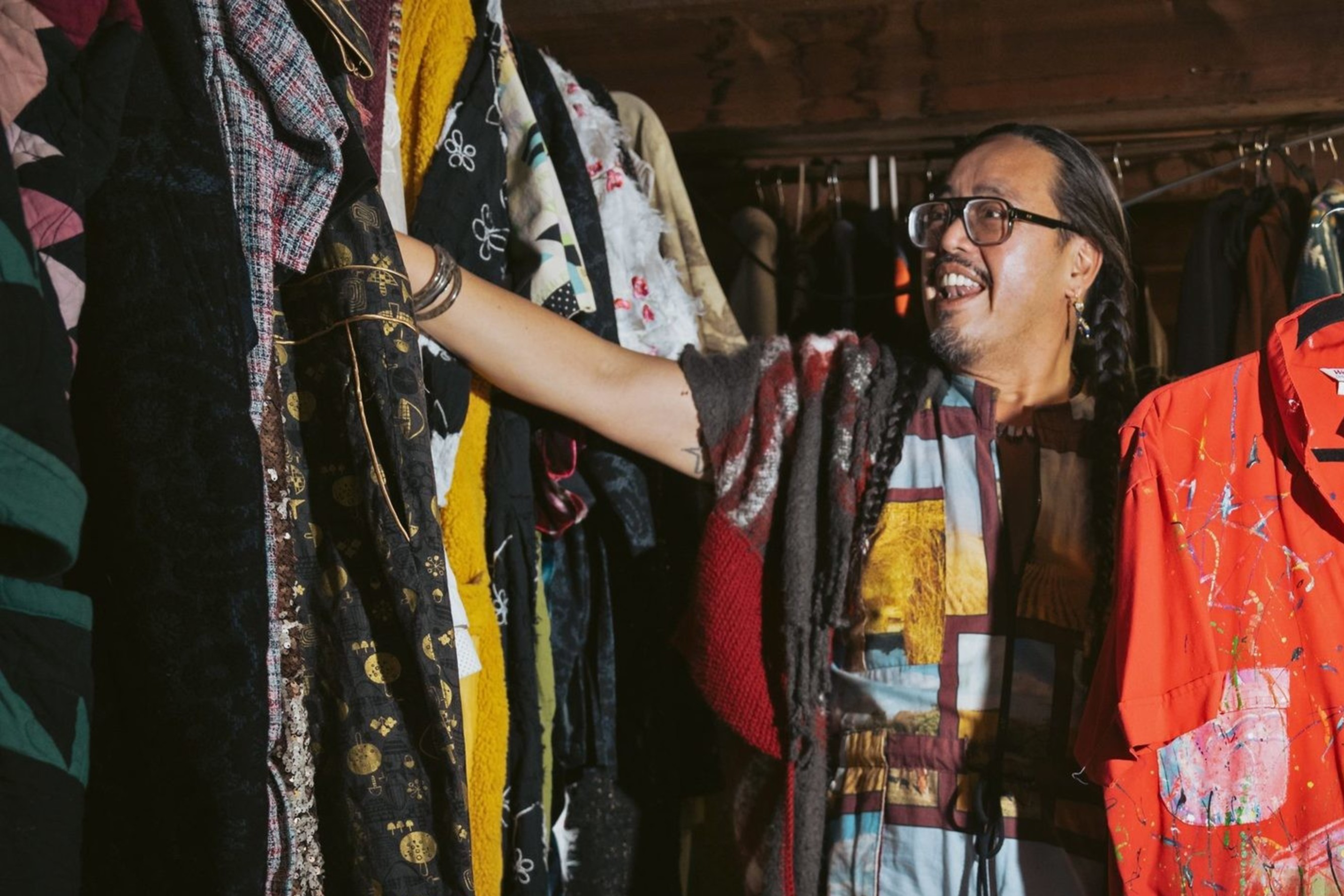 A person stands in a room filled with colorful clothing on racks. They are holding a vivid red jumpsuit splattered with paint. The space is warmly lit and eclectic.