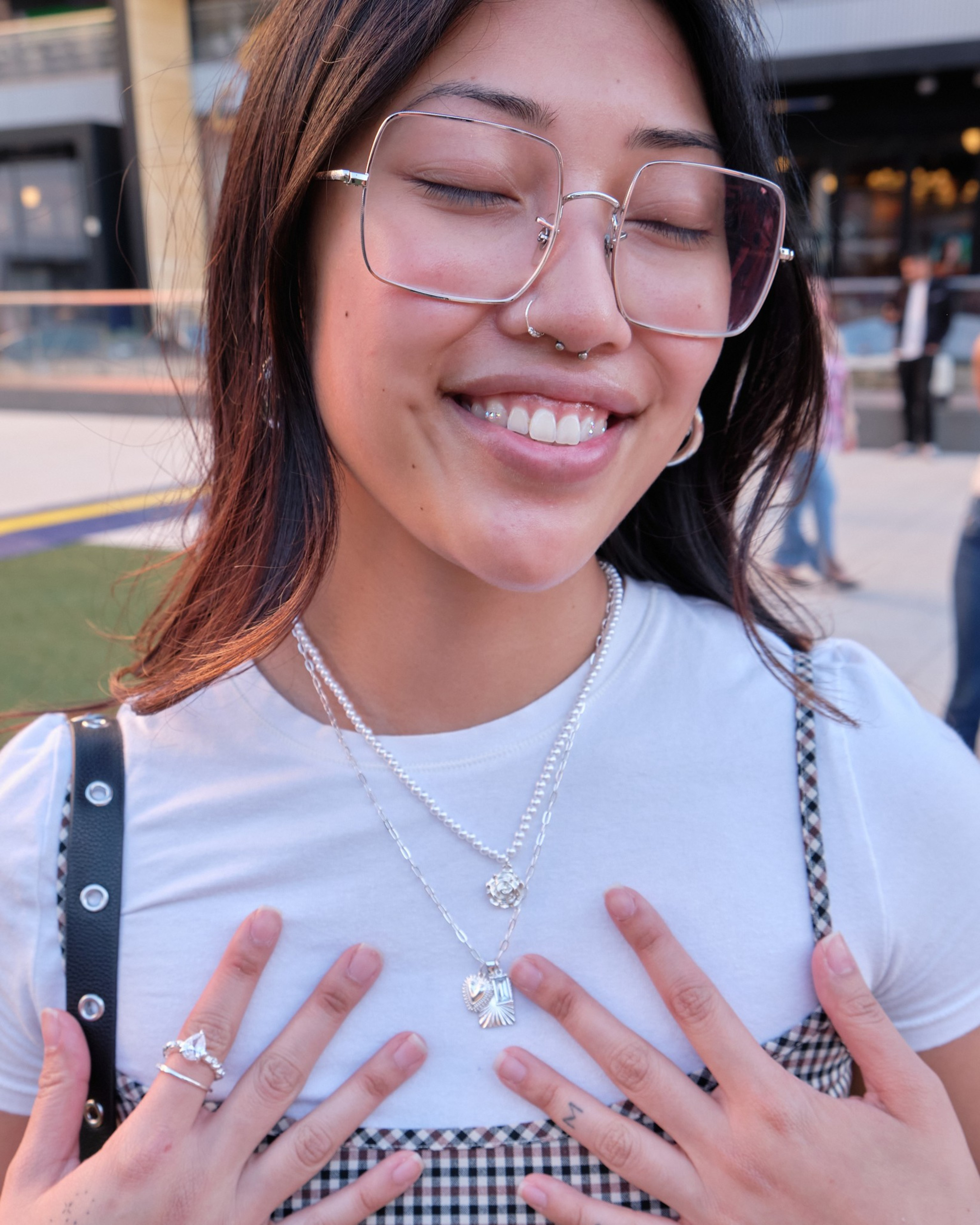 A woman with closed eyes smiles, wearing large glasses, multiple necklaces, rings, and a checkered top over a white shirt, with her hands near her chest.