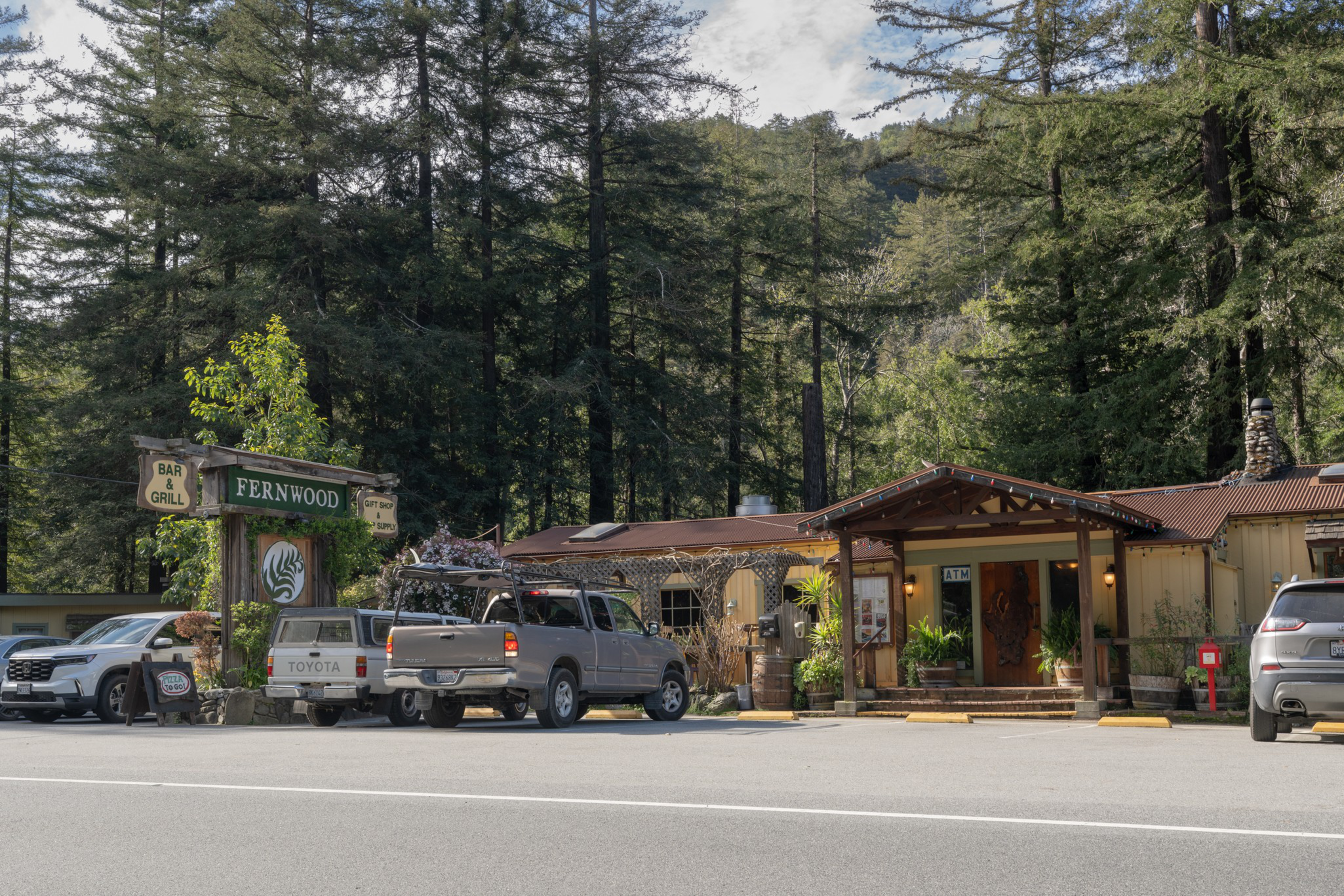A rustic bar and grill named Fernwood sits by a roadside, surrounded by tall pine trees, with several parked vehicles nearby.