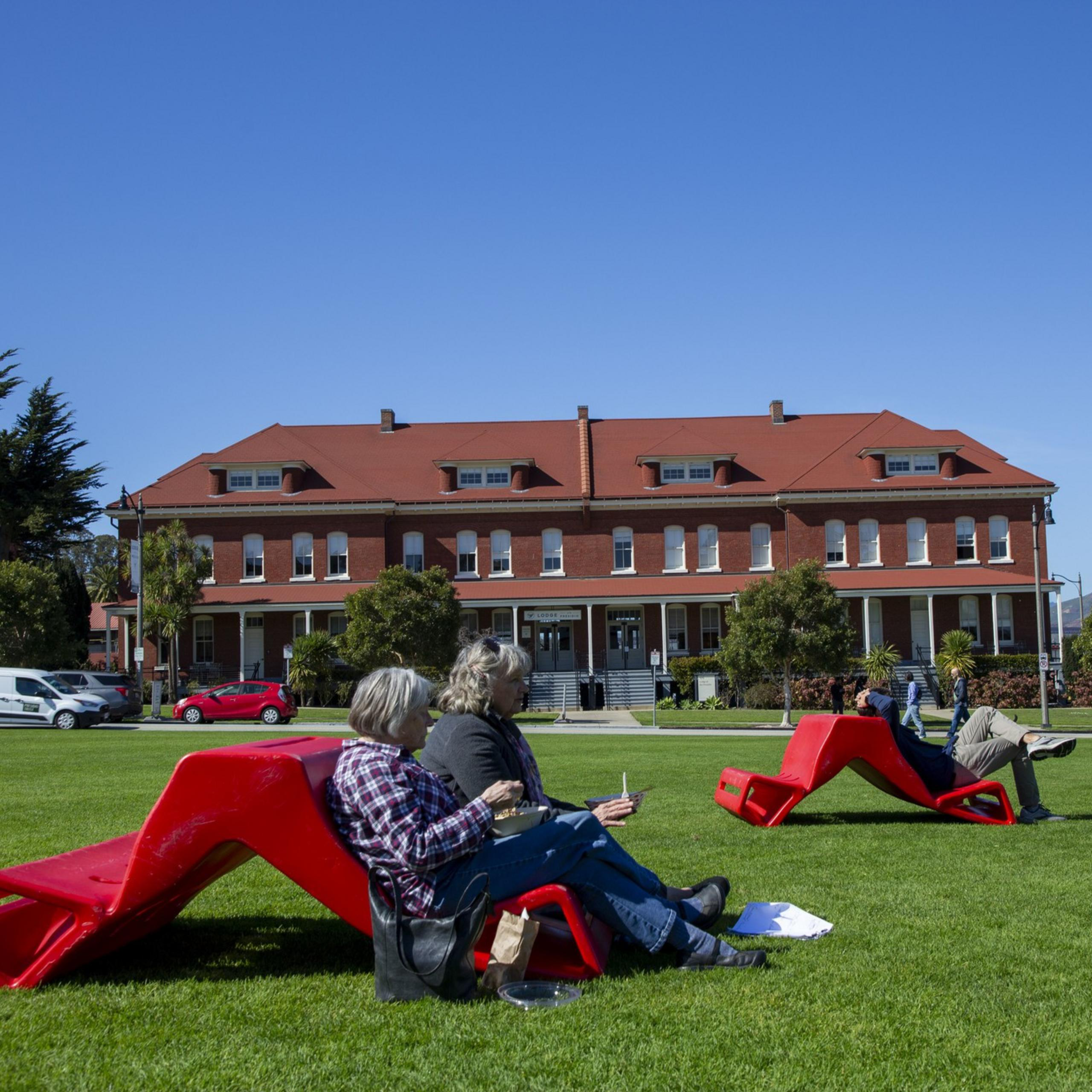 People are sitting on red lounge chairs on a grassy lawn in front of a large, red-roofed brick building under a clear blue sky.