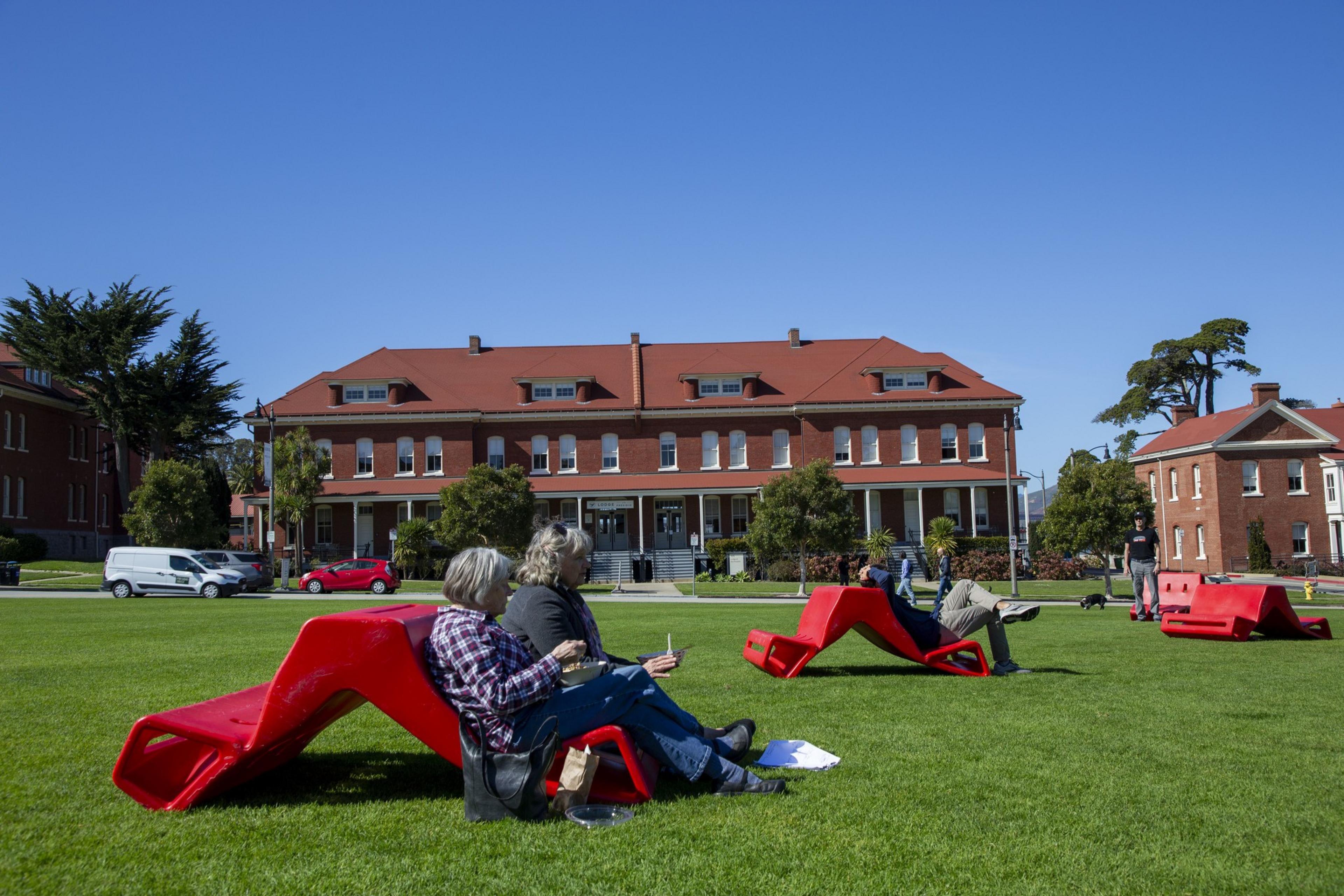 People are sitting on red lounge chairs on a grassy lawn in front of a large, red-roofed brick building under a clear blue sky.