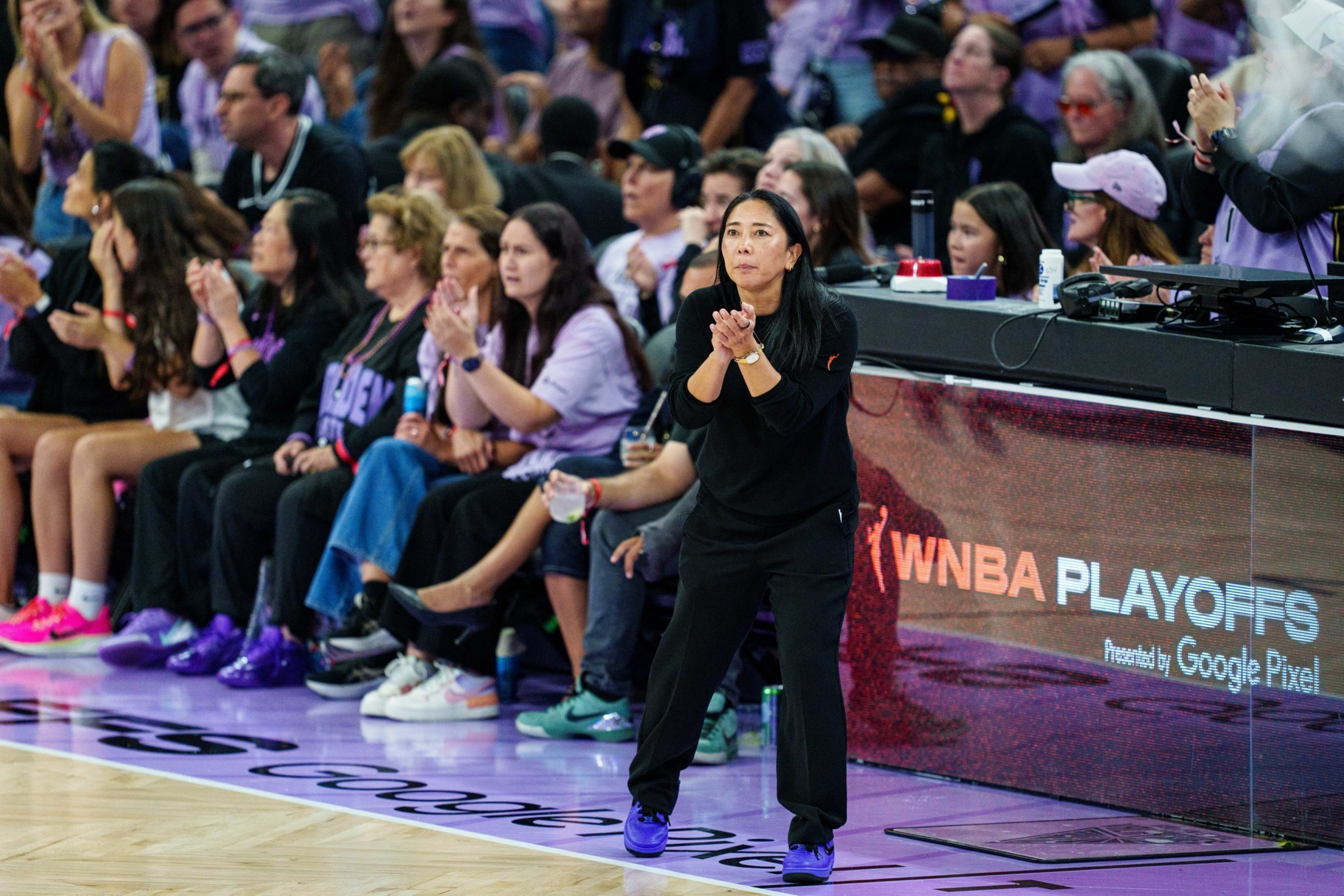 A woman in black sportswear claps on the sidelines of a basketball court during a WNBA Playoffs game with seated spectators behind her.