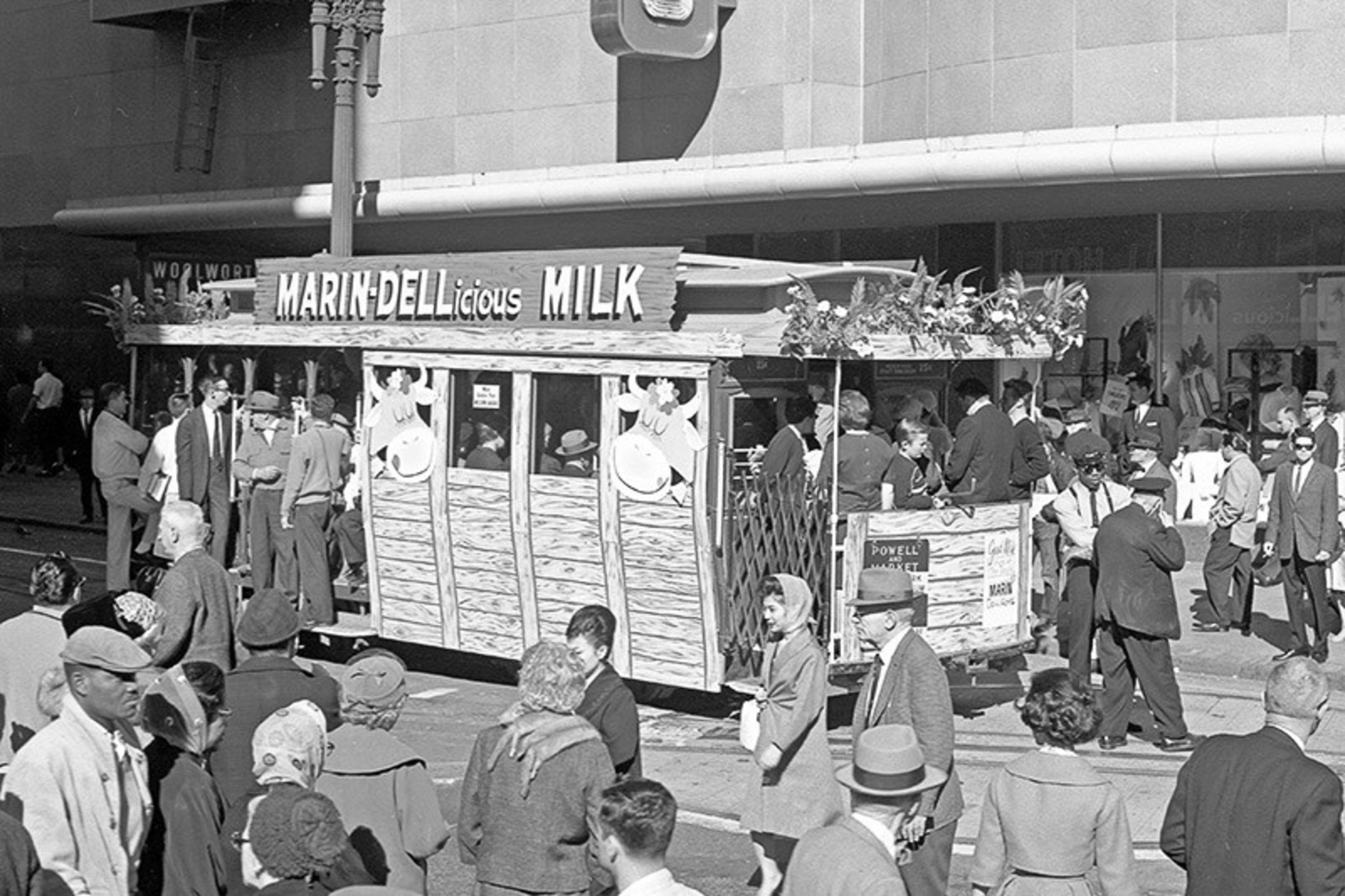 A crowded street scene with a wooden trolley advertising "MARIN-DELLicious MILK" surrounded by people dressed in mid-20th century attire.