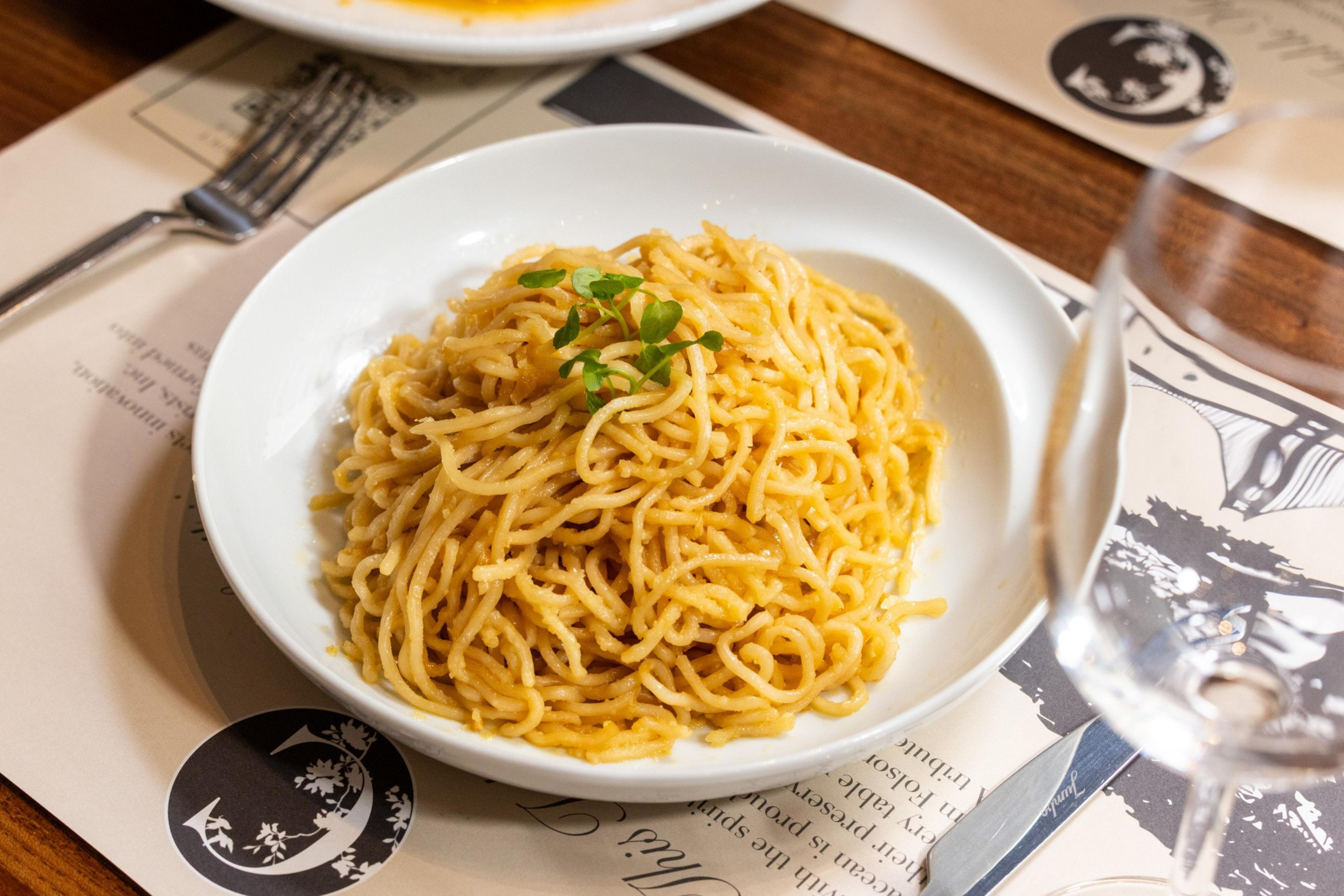 A white bowl filled with spaghetti noodles garnished with a small sprig of green herbs sits on a placemat next to a fork and an empty wine glass.