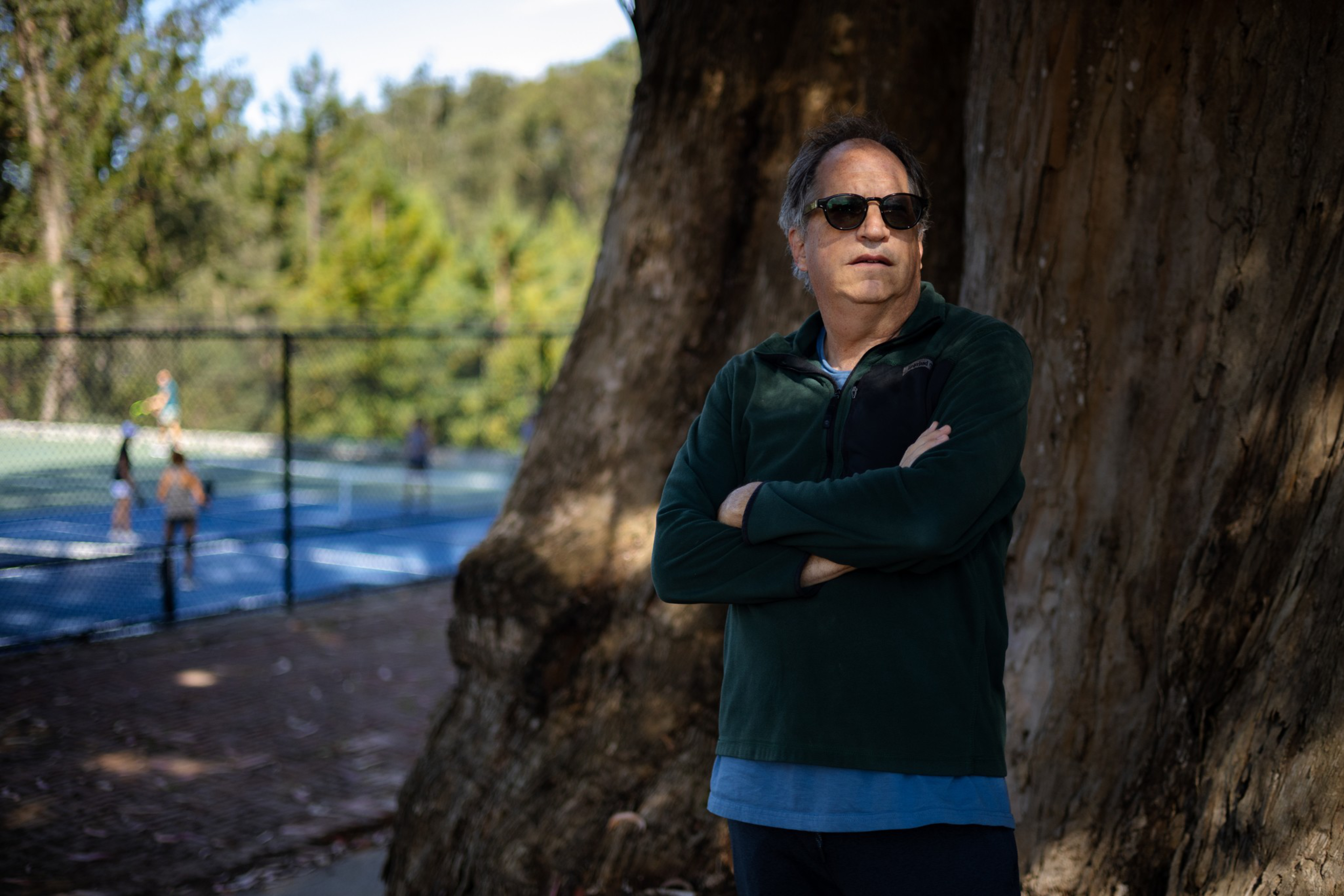 A man wearing sunglasses and a dark green jacket stands with arms crossed leaning against a large tree, with people playing tennis on a court in the background.