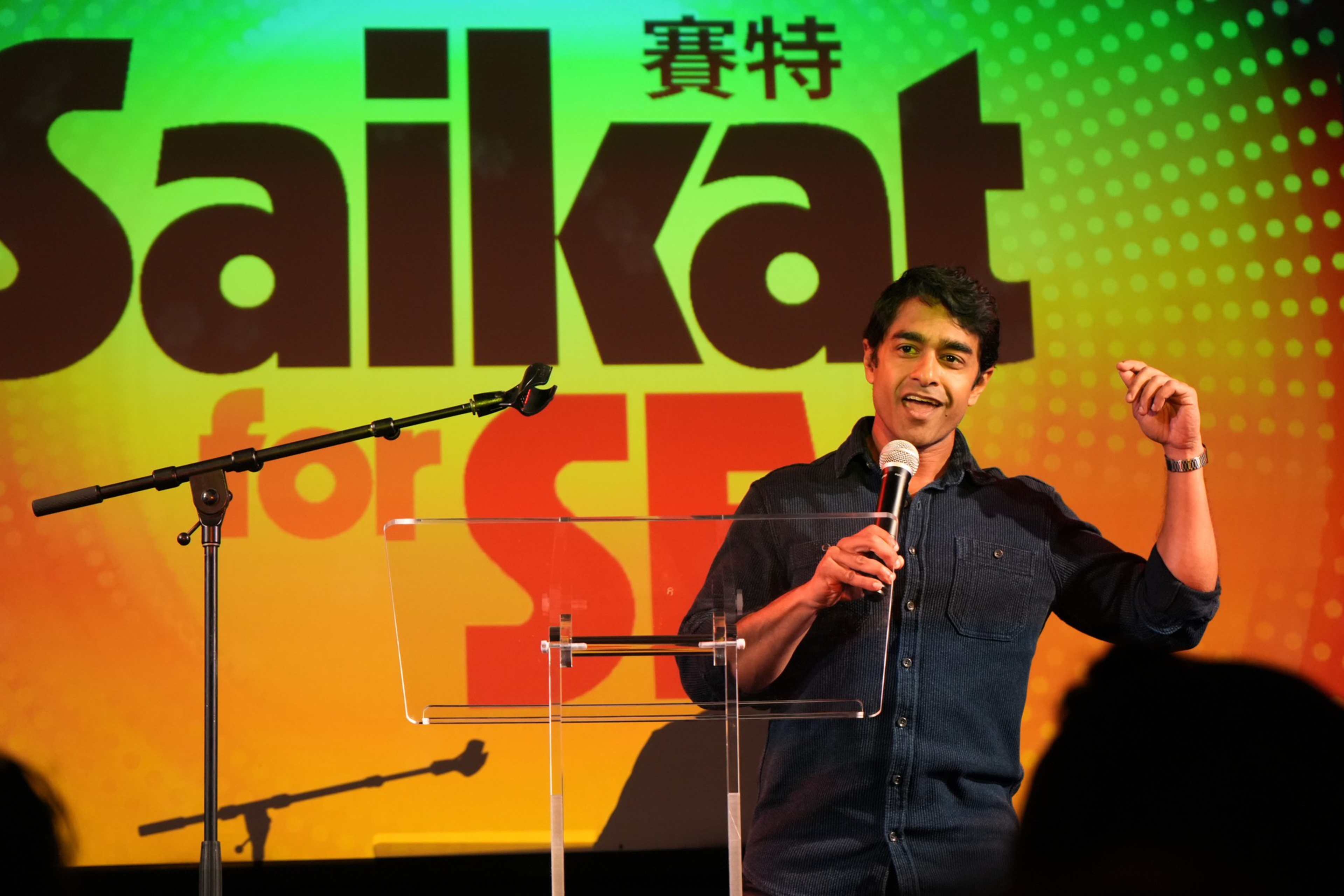 A man in a dark shirt speaks into a microphone, standing behind a clear podium with a colorful “Saikat for SE” backdrop behind him.