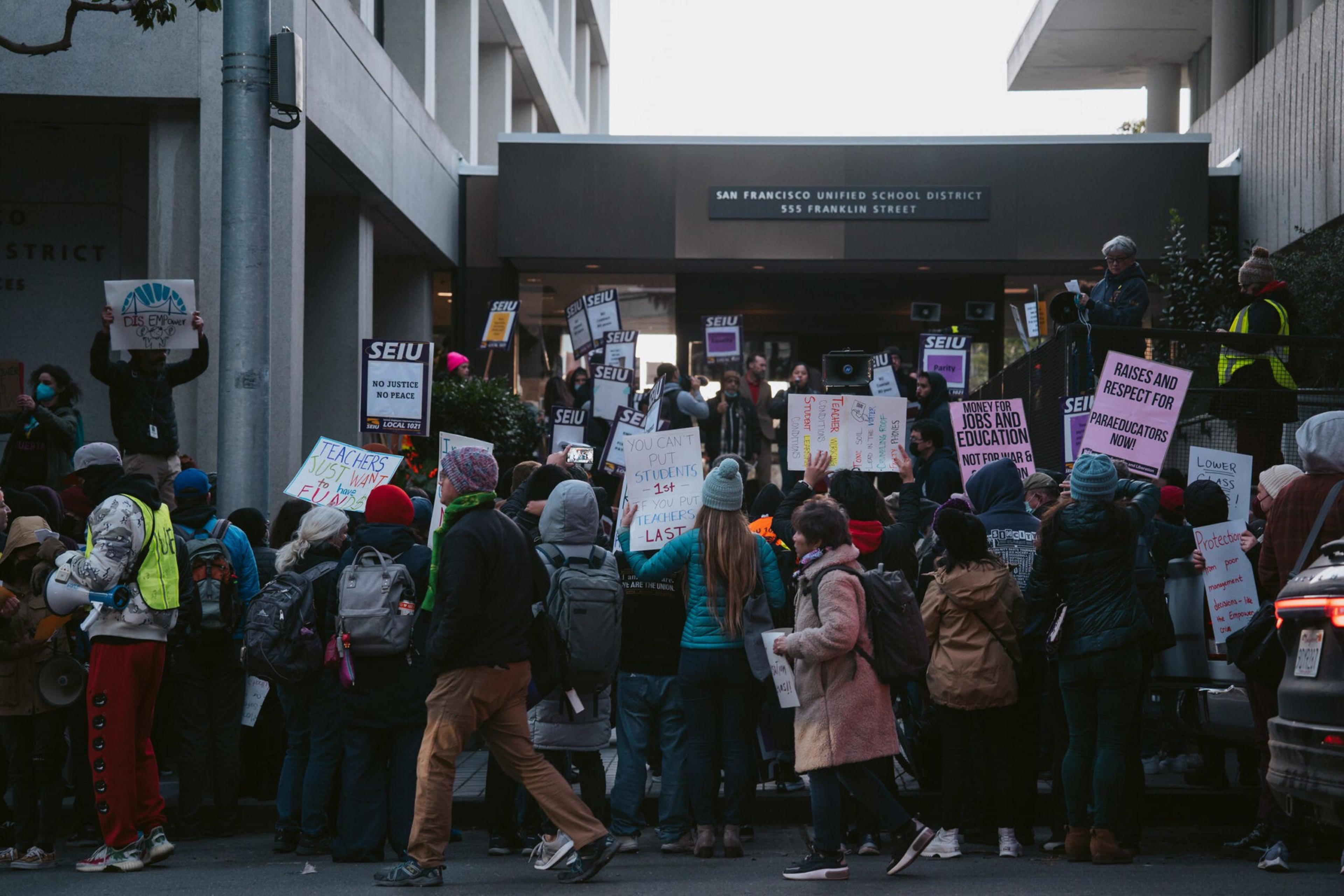 A crowd gathers outside a building holding signs demanding justice, respect, fair wages, and support for teachers and school staff.