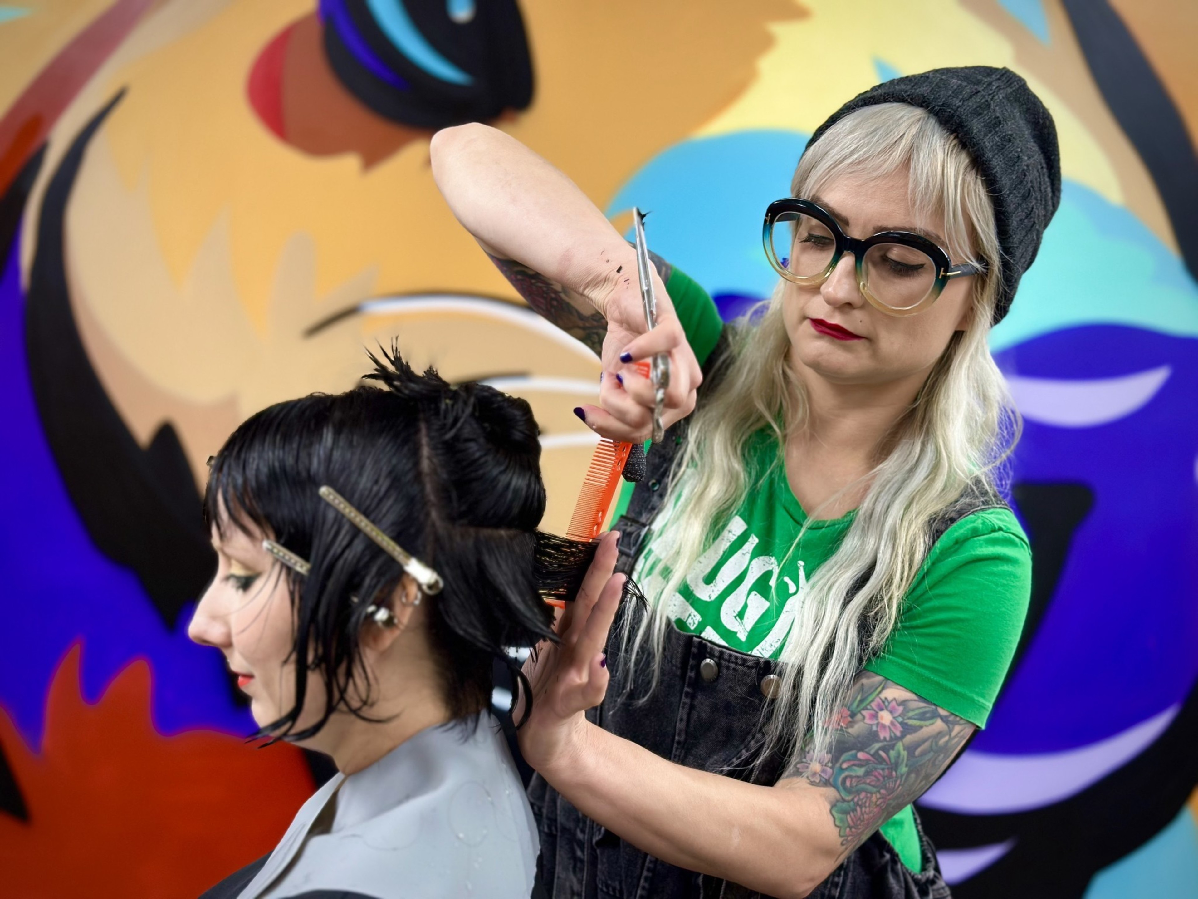 A hairstylist with long blonde hair, large glasses, a beanie, and tattoos cuts a woman's short black hair with scissors against a colorful, abstract mural backdrop.