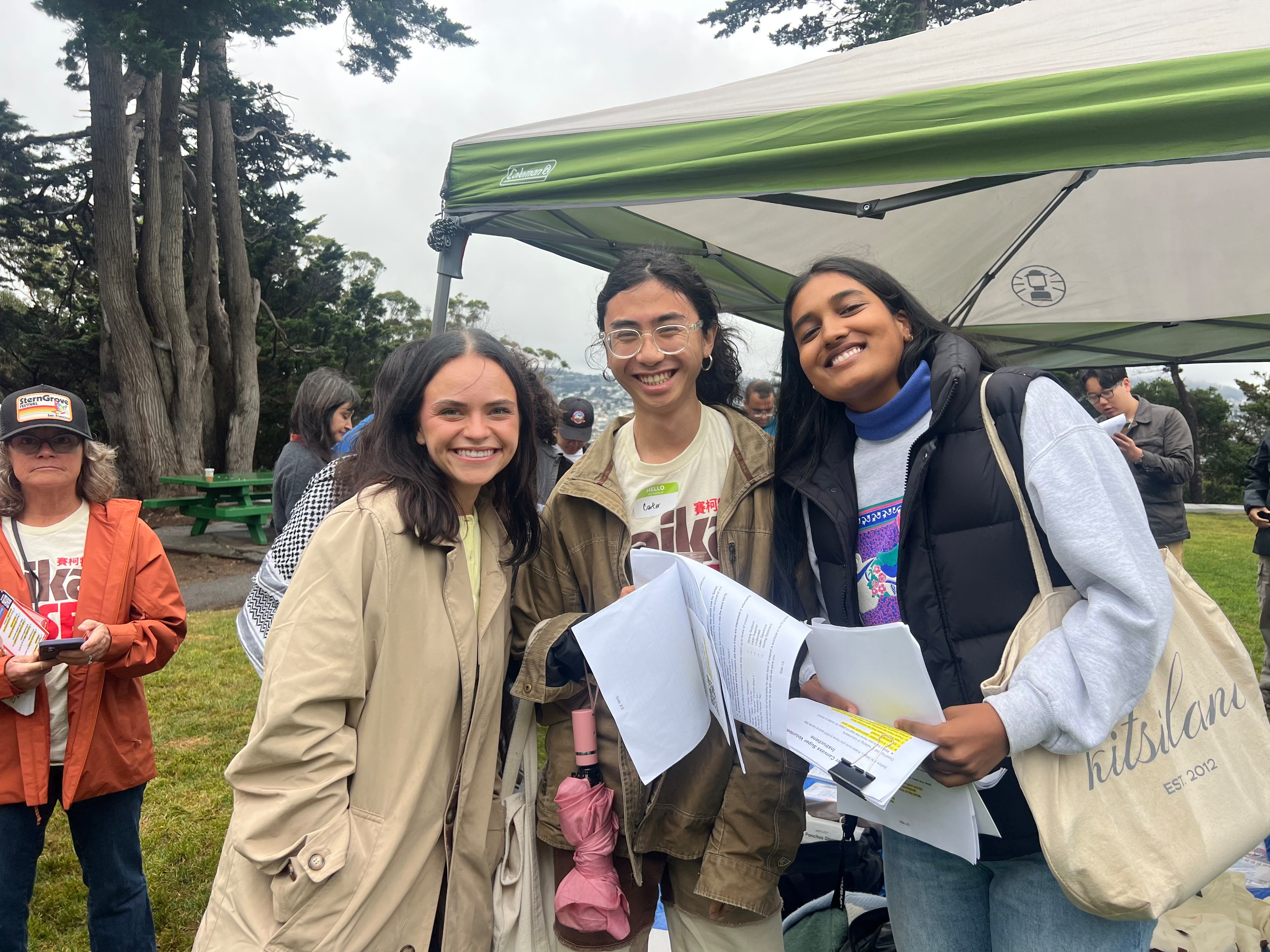Three smiling young people hold papers under a green canopy at an outdoor event with trees and others in the background.