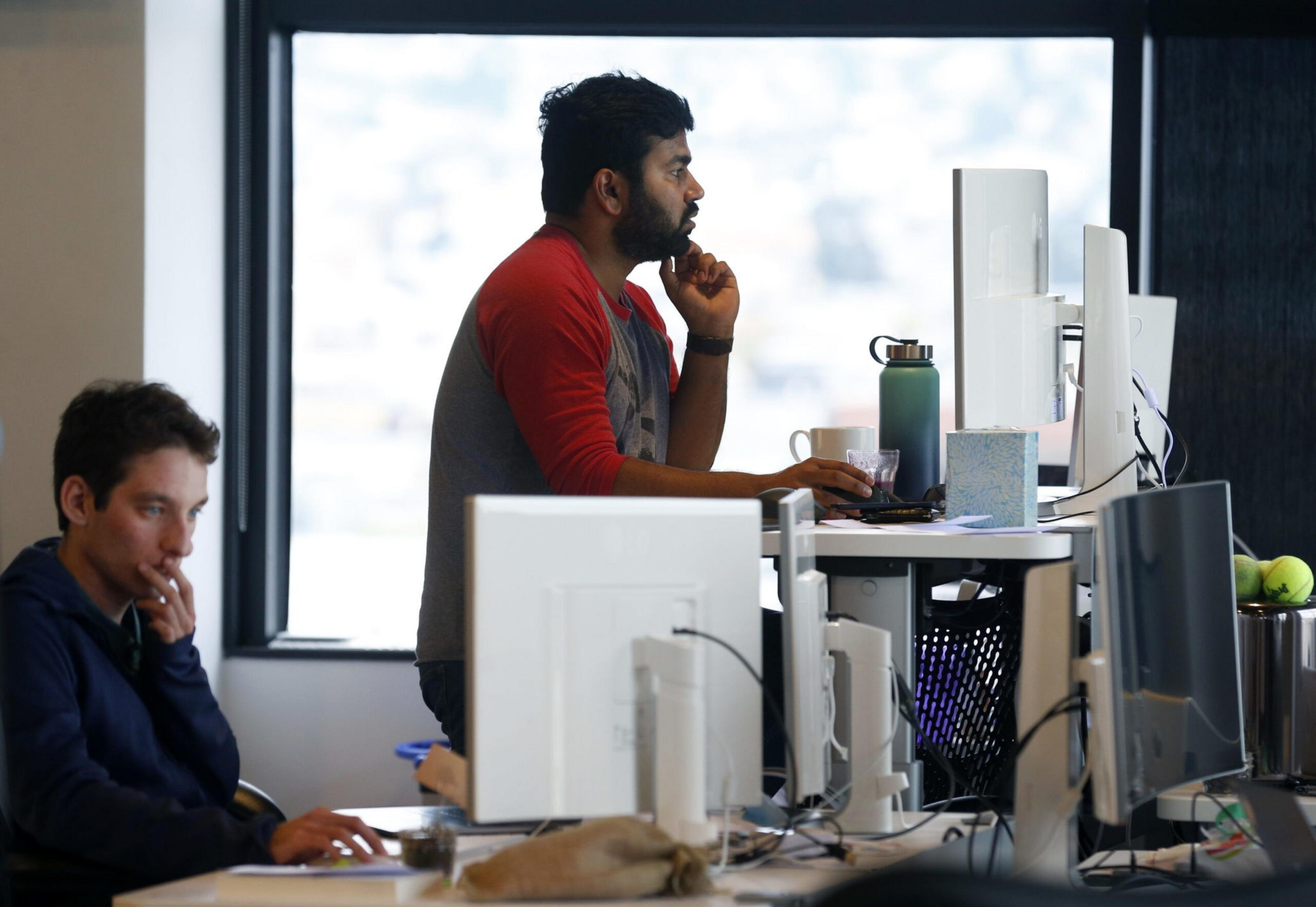 A man in a red and gray shirt stands thoughtfully at a desk with a computer, while another man sits nearby focused on his work.