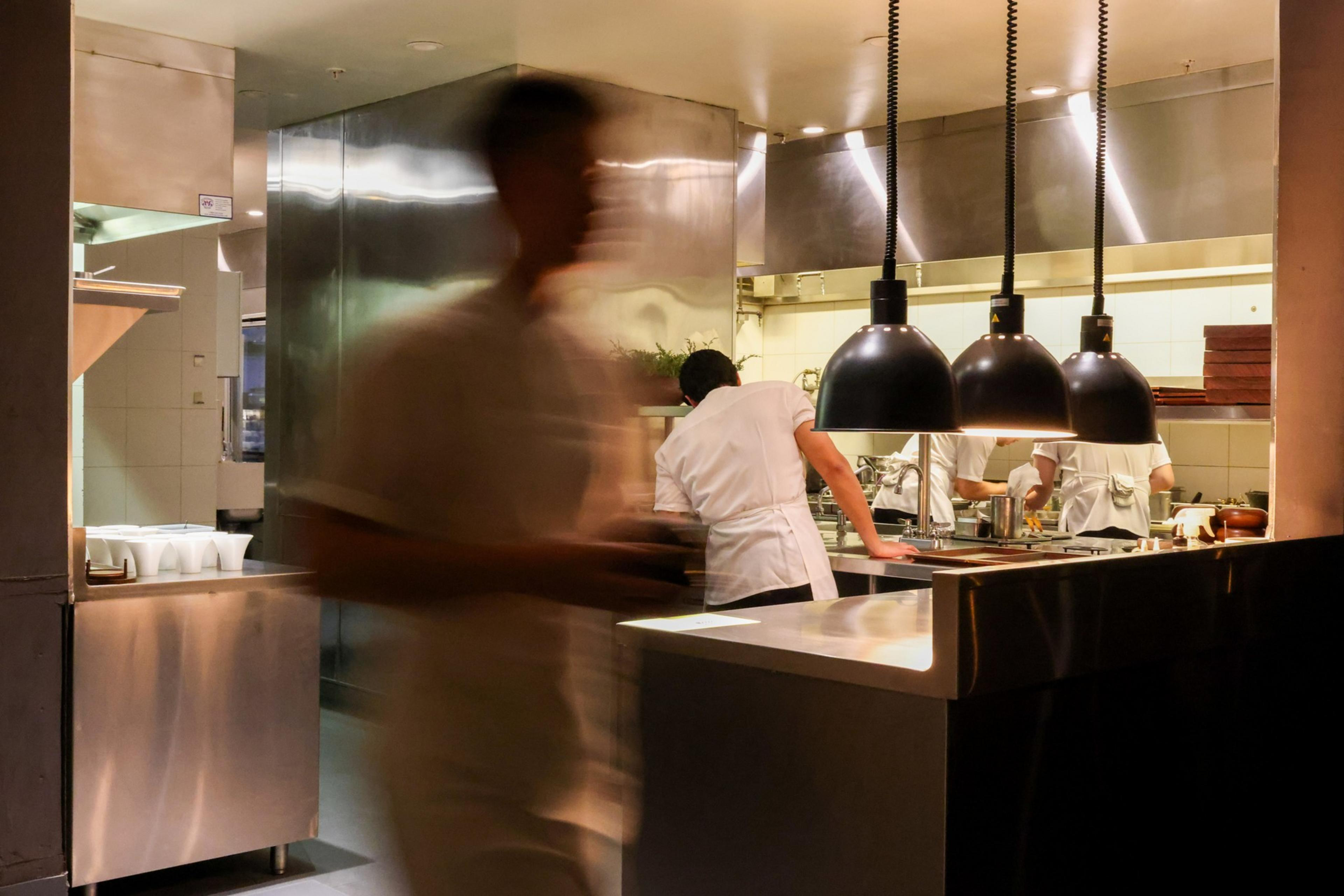 A busy restaurant kitchen shows chefs in white uniforms working efficiently. The lighting is warm, featuring industrial pendant lights, with a blurred figure moving quickly.