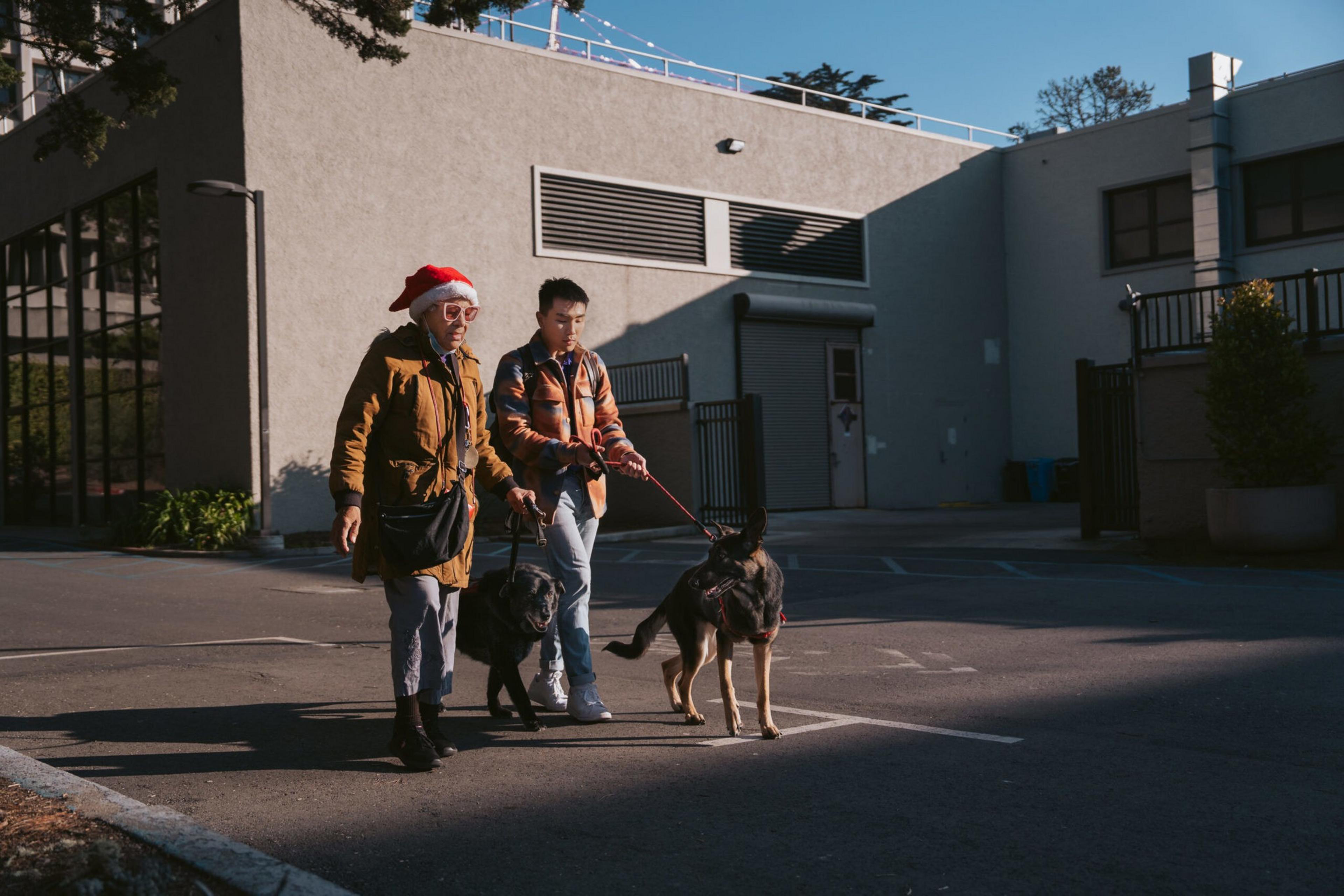 Two people walk dogs on leashes in a parking lot beside a building, with one person wearing a Santa hat and both dressed in jackets.