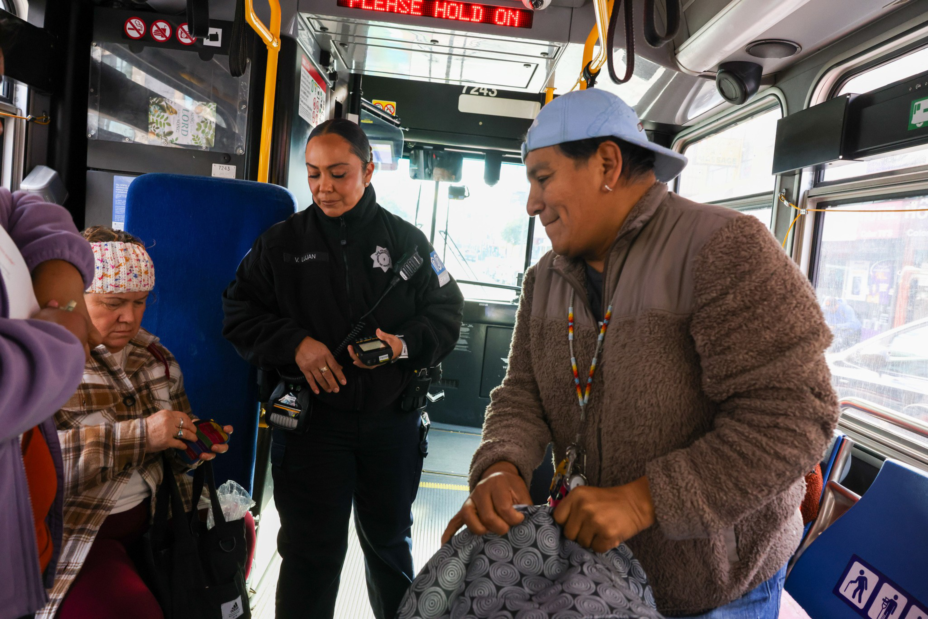 A bus interior shows a police officer speaking with a seated woman holding a card. Nearby, a man in a furry jacket and baseball cap grips a patterned bag.