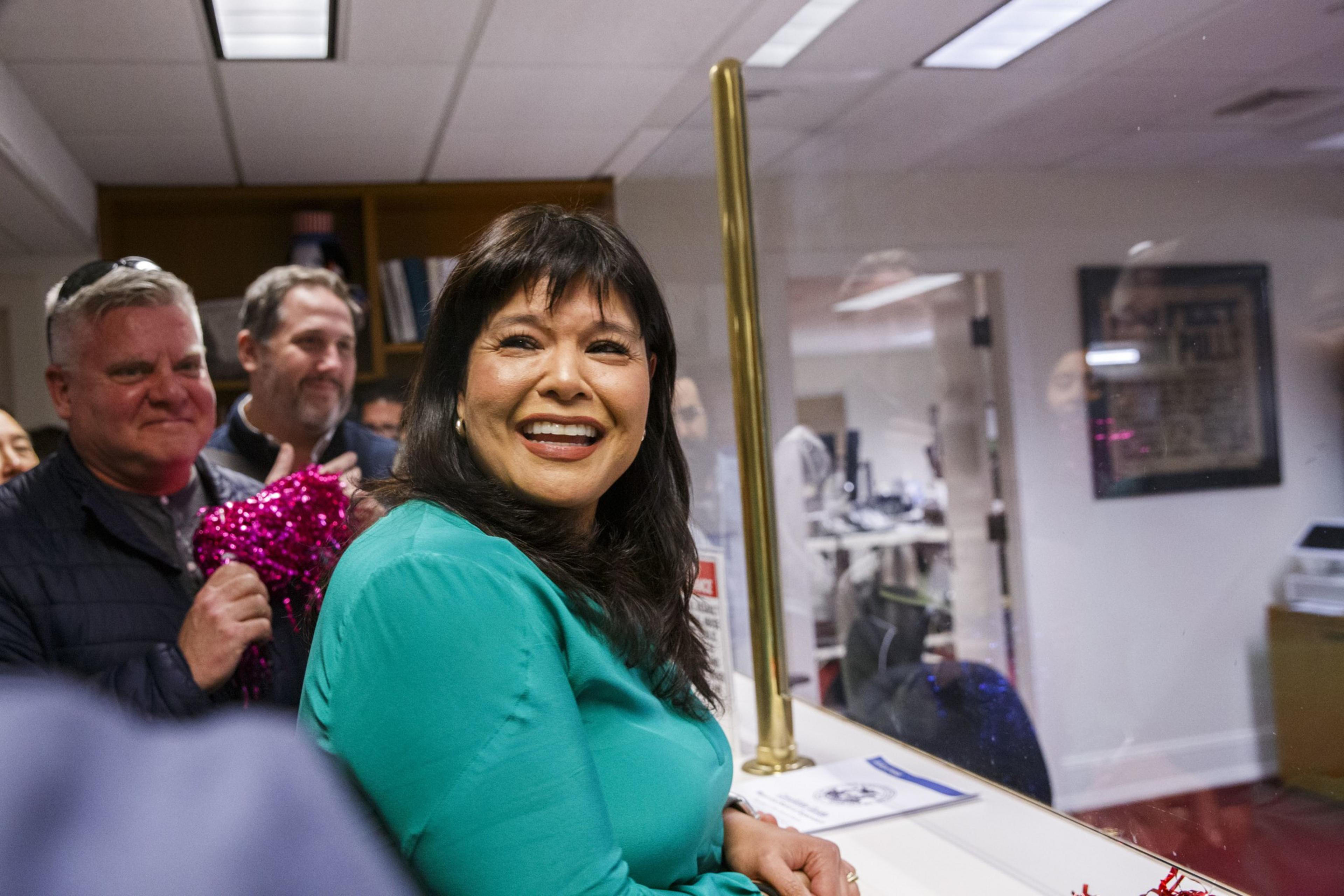 A smiling woman in teal attire is foregrounded with onlookers behind her in an indoor setting.