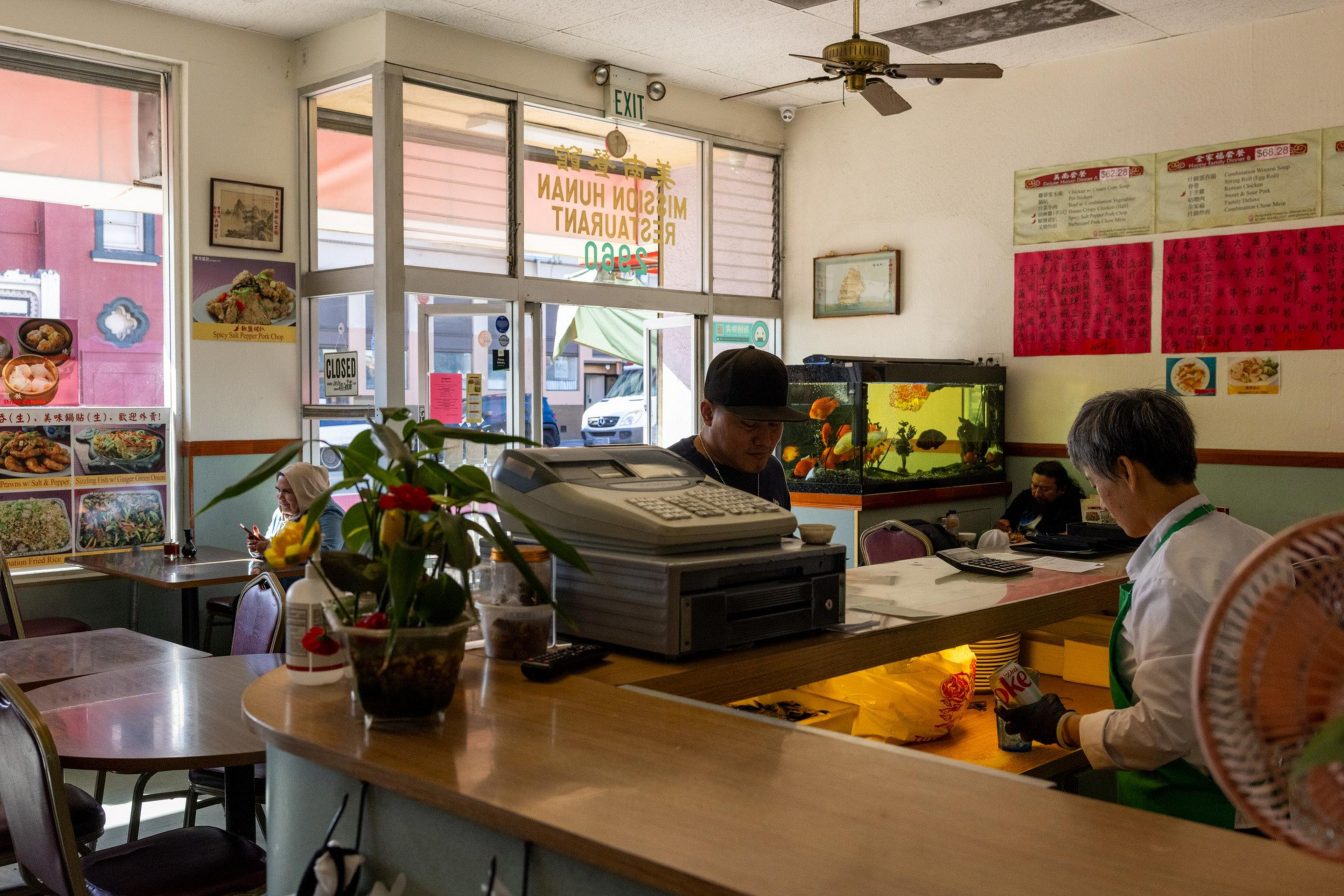A small restaurant interior features customers seated by windows, a man and woman behind the counter, an aquarium, and menus on the walls.