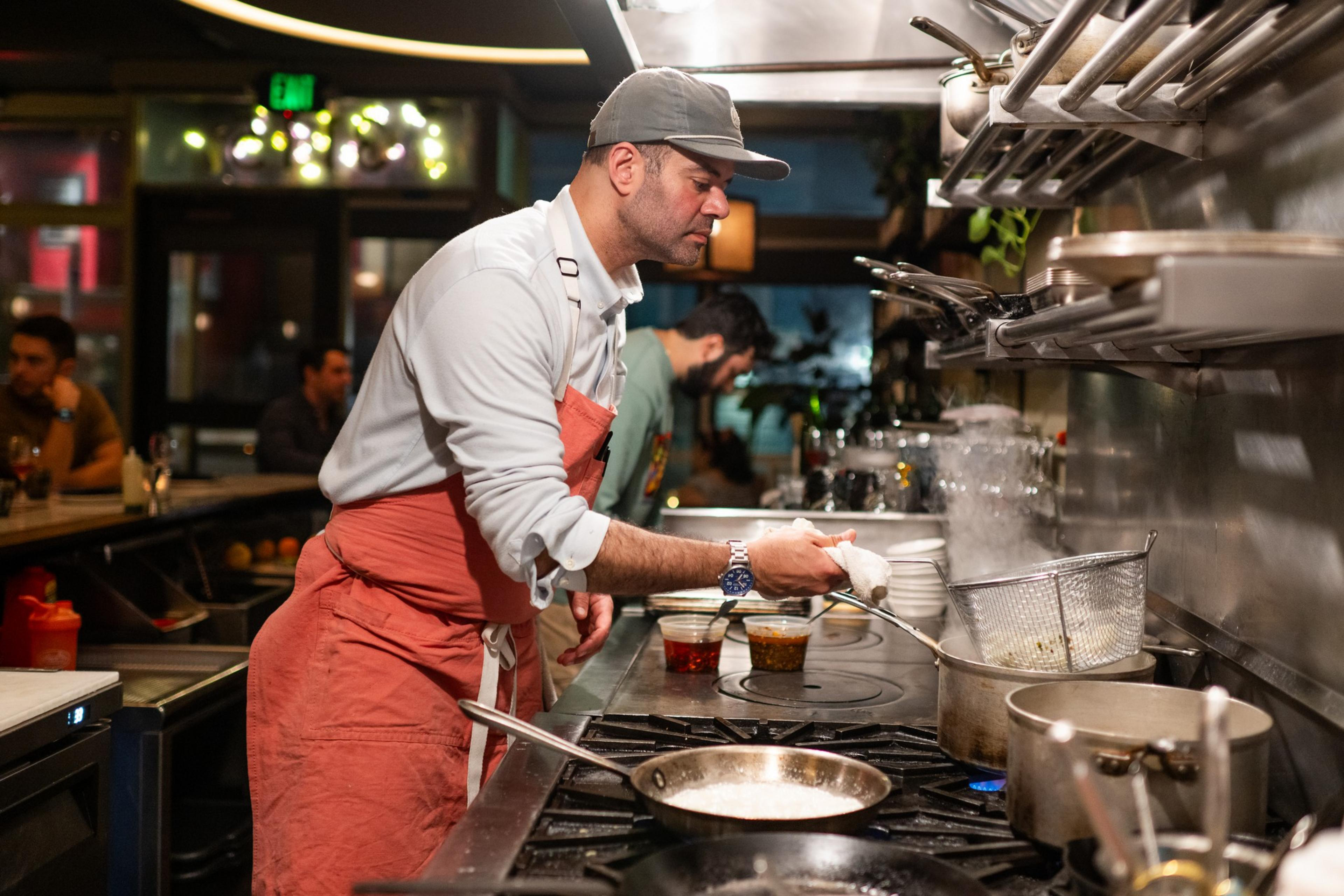 A chef in a pink apron and cap is cooking in a restaurant kitchen, tending to a steaming pot on the stove. The background shows a lively dining area.