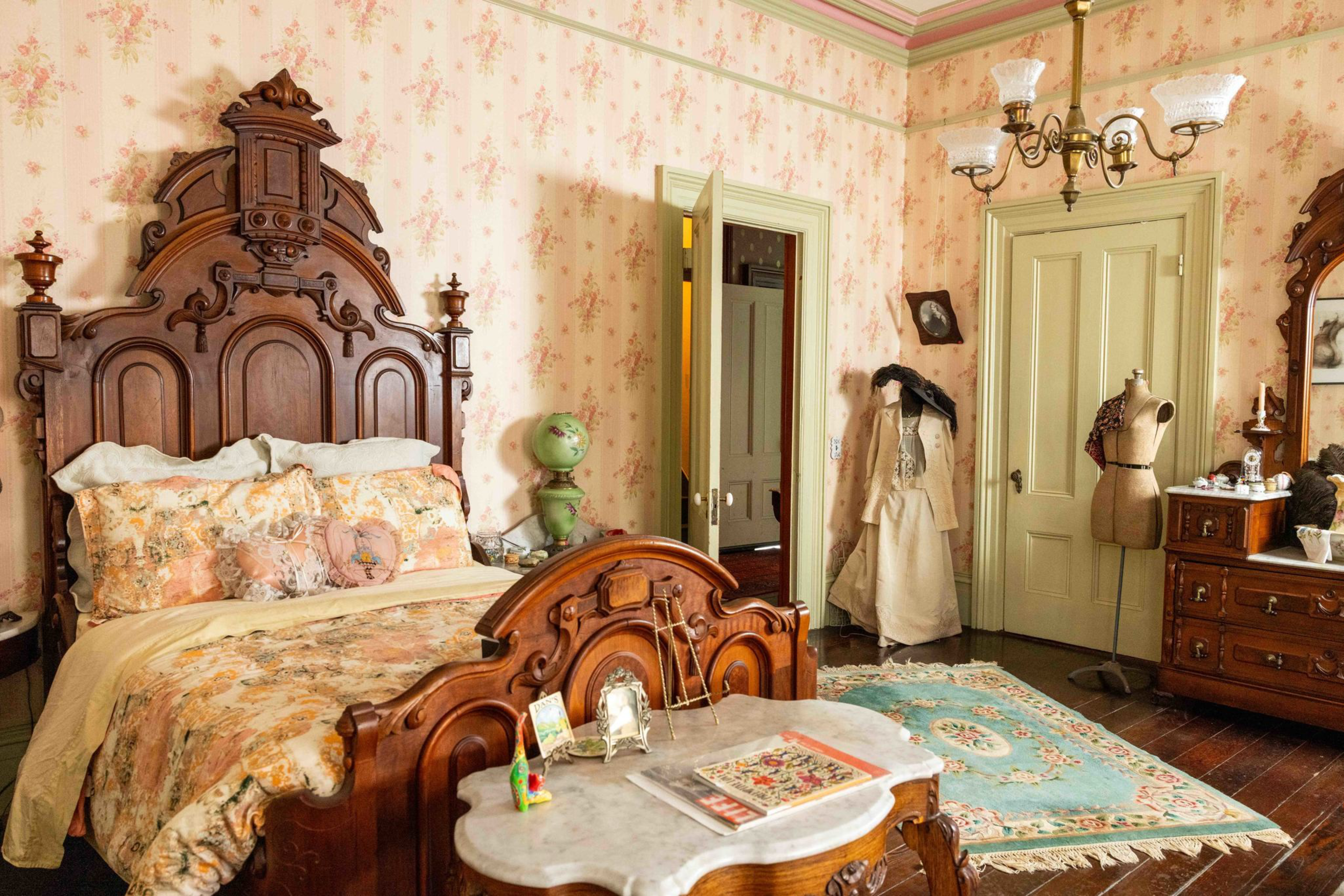 A vintage bedroom with ornate wooden bed, floral bedding, Victorian dress, dress form, wooden dresser with mirror, beige doors, wallpaper, and a floral rug.