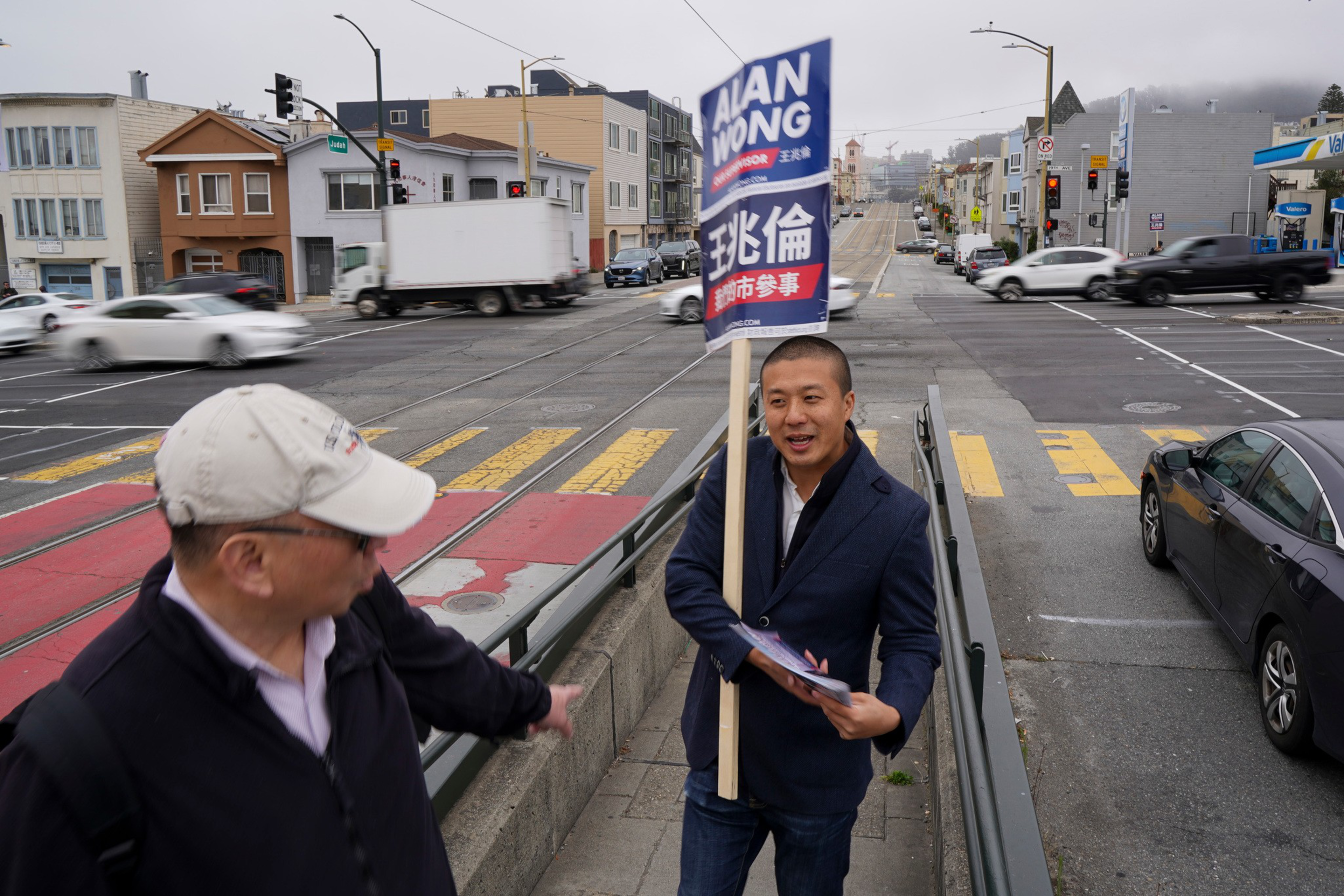 A man holding a campaign sign and flyers talks to an older man wearing a baseball cap at a busy city street intersection.