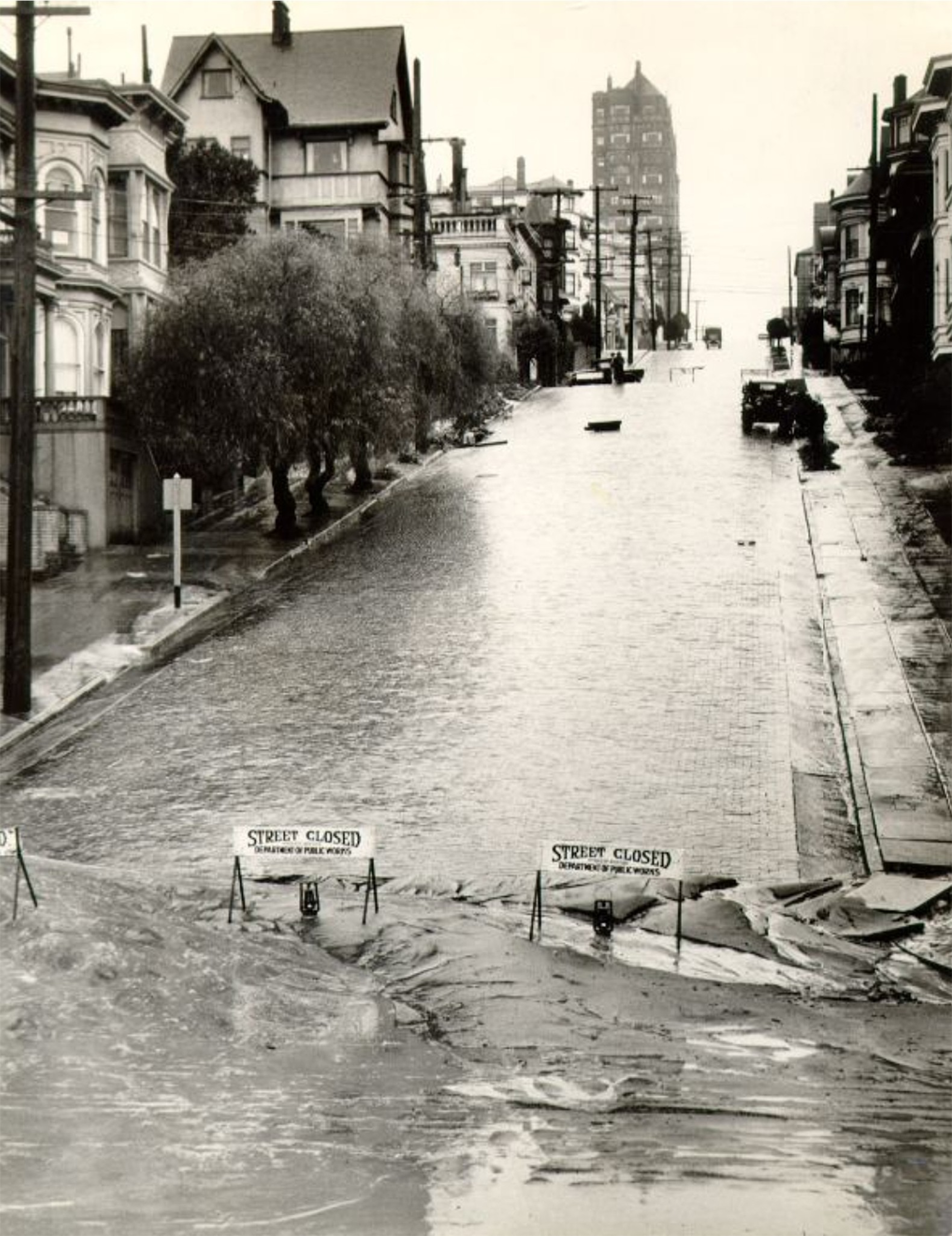 A flooded street with mud and water blocking it near the bottom, flanked by trees and buildings, with "Street Closed" signs placed across the road.