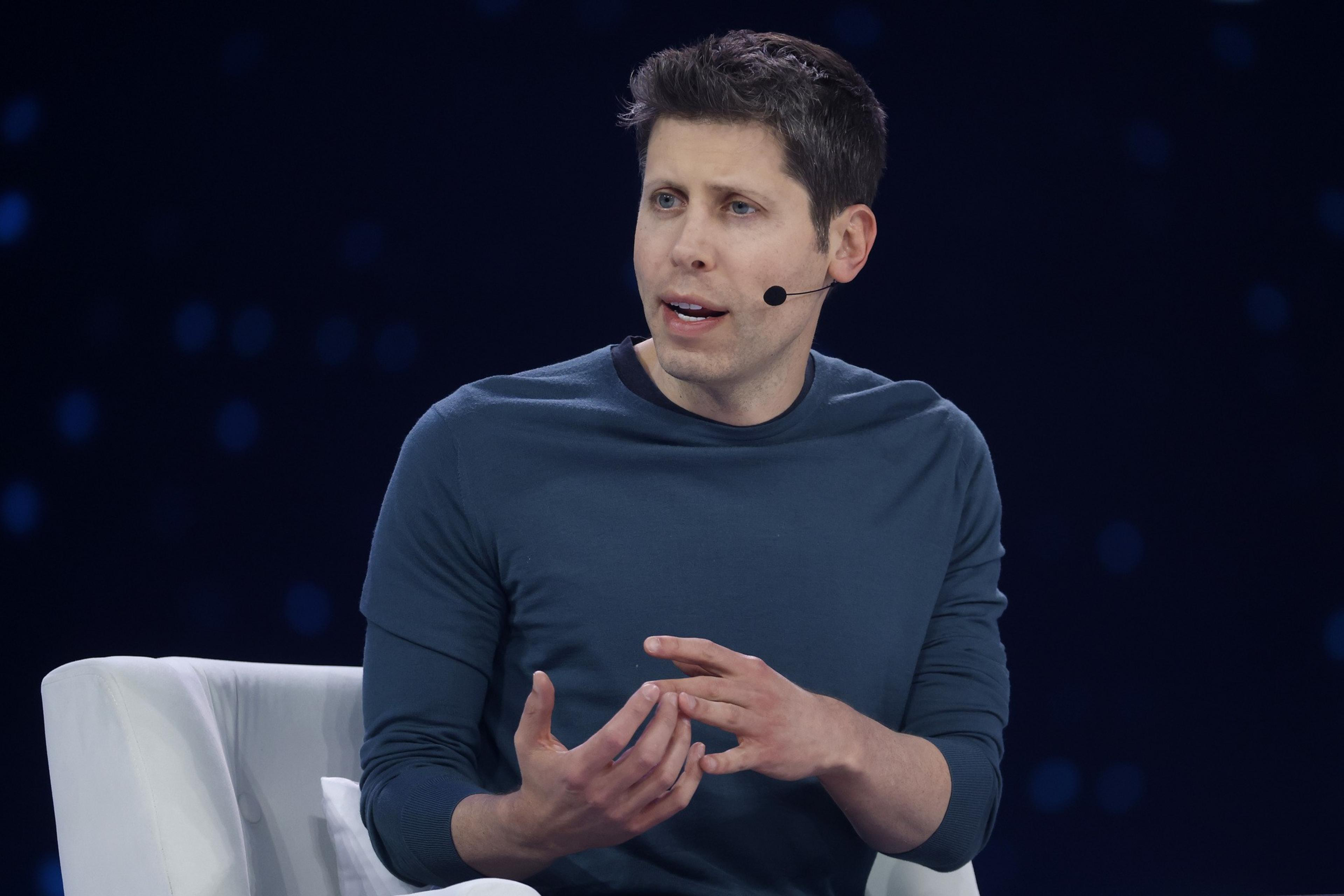 A man wearing a dark blue long-sleeve shirt sits in a white chair, speaking with hand gestures and wearing a headset microphone.