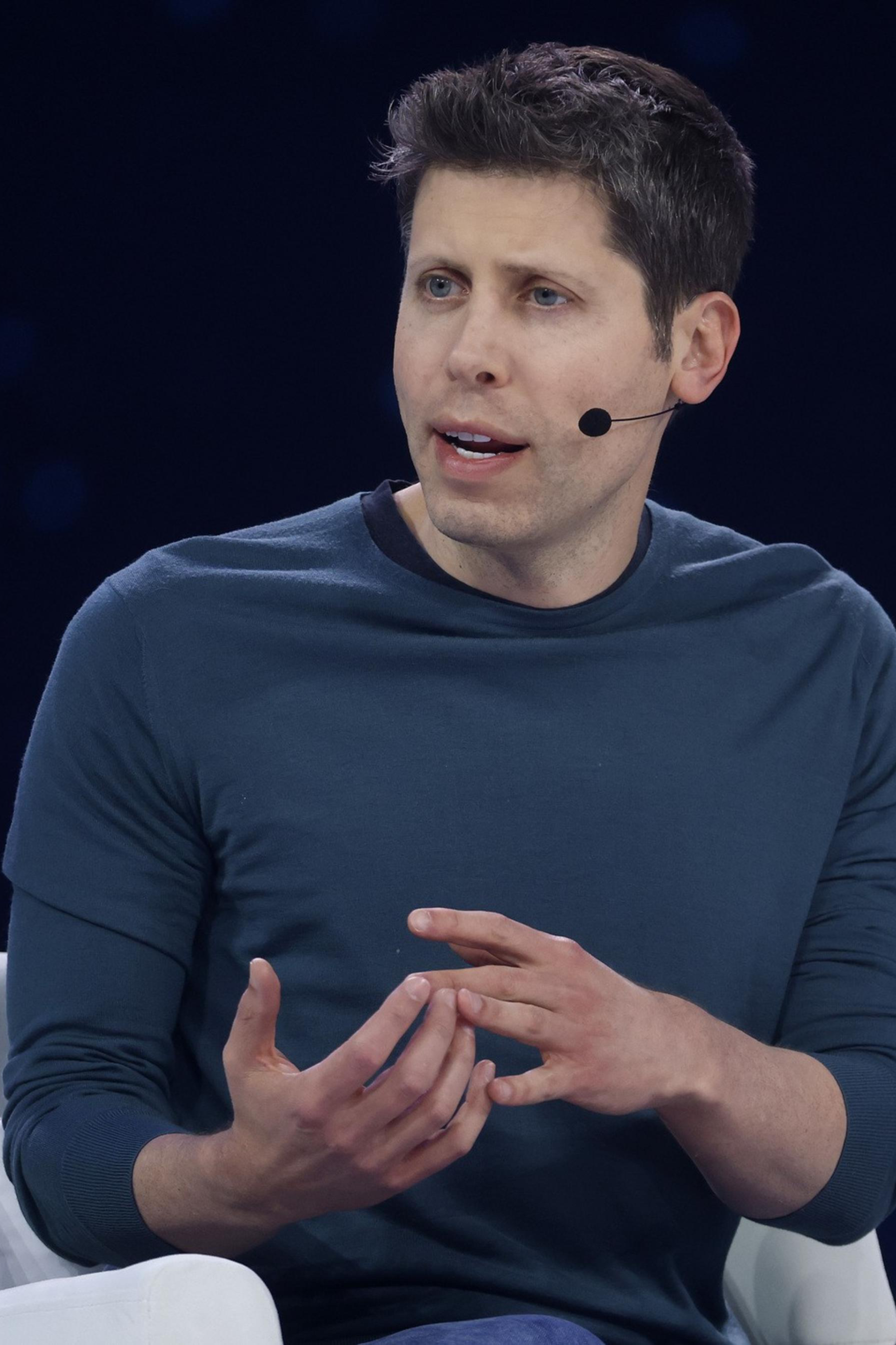 A man wearing a dark blue long-sleeve shirt sits in a white chair, speaking with hand gestures and wearing a headset microphone.