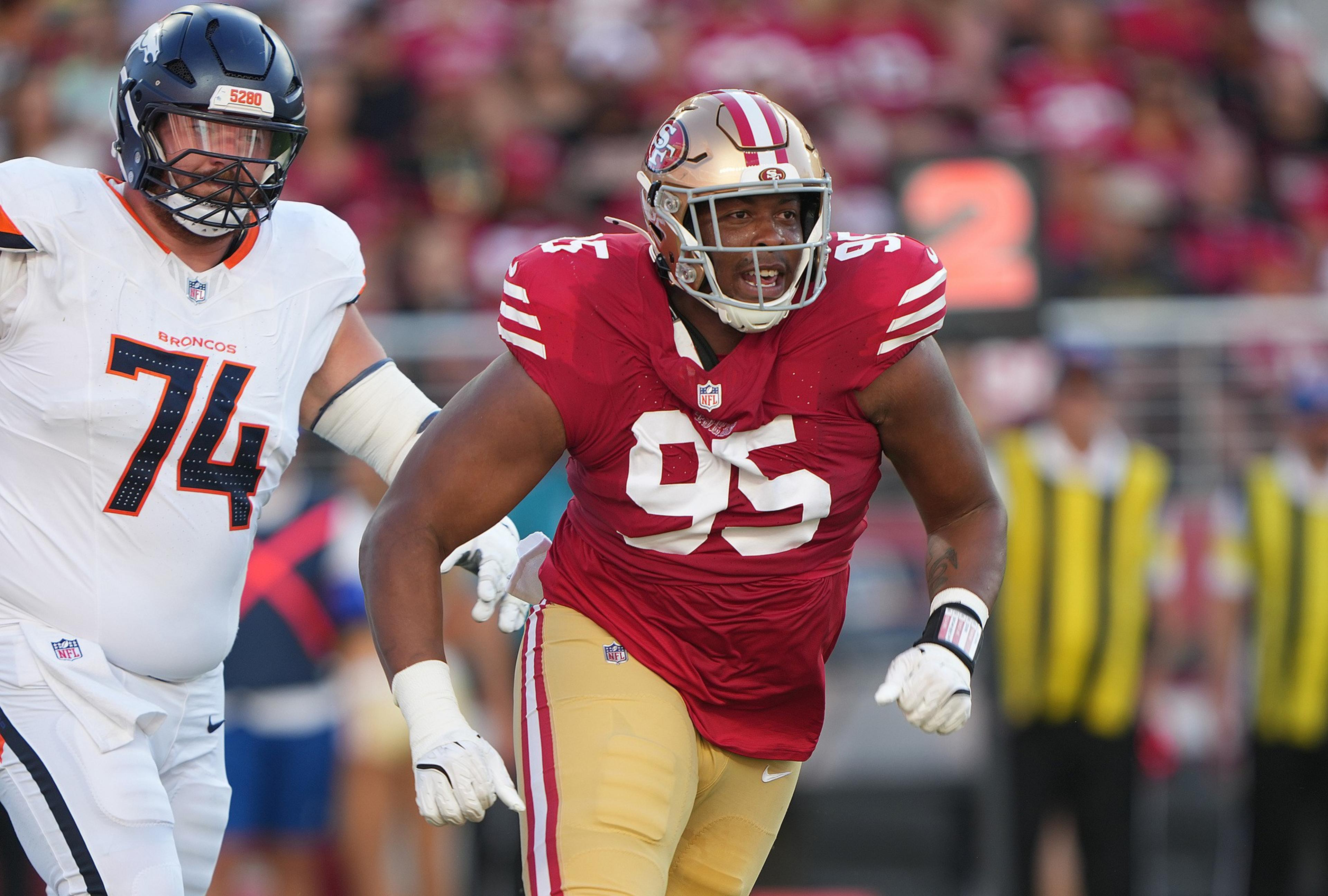 A football player in a red and gold 49ers uniform, number 95, is running past a Denver Broncos player wearing a white jersey, number 74.