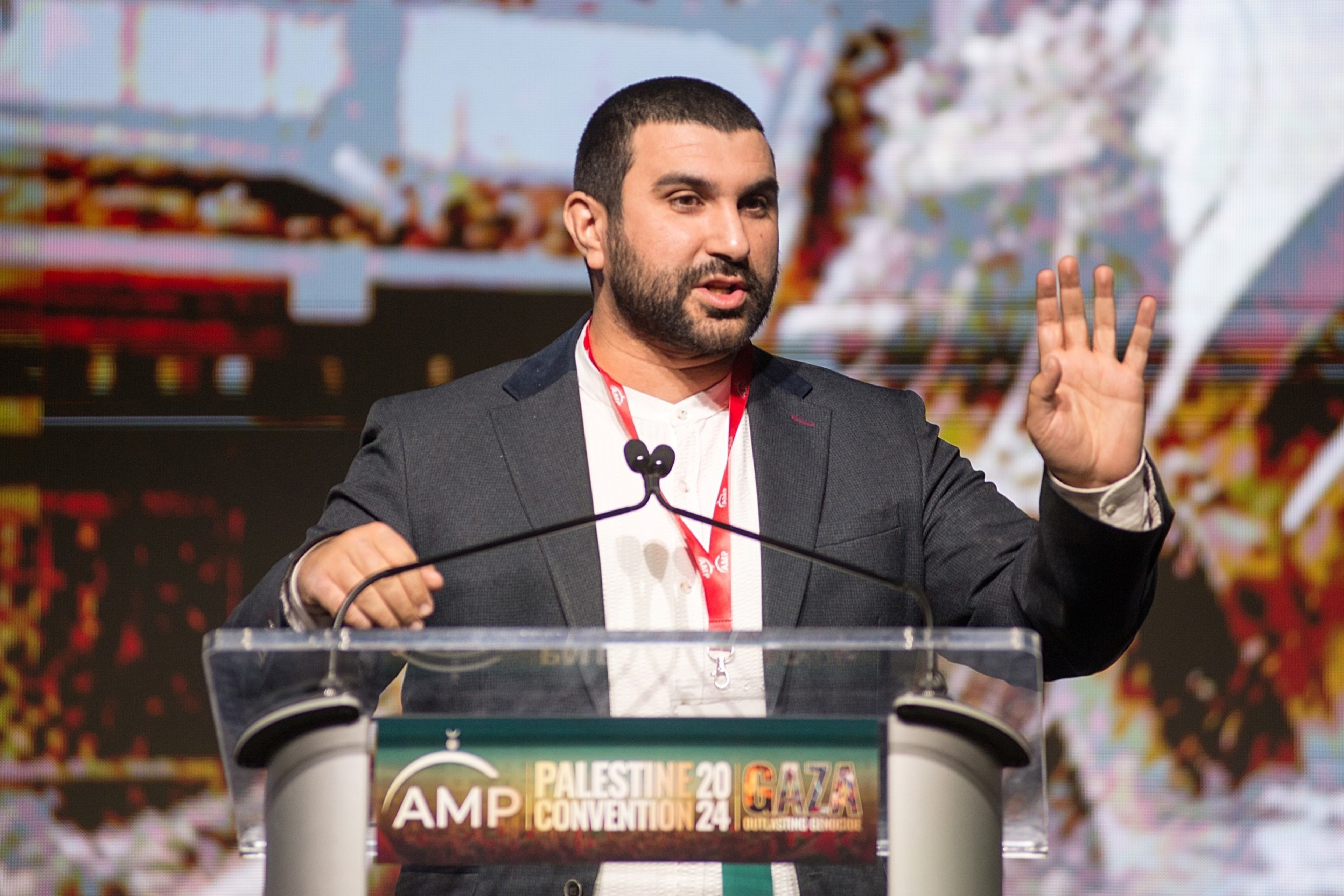 A man wearing a dark blazer and white shirt speaks at a podium with a red lanyard, gesturing with one hand raised during the AMP Palestine 2024 Convention.