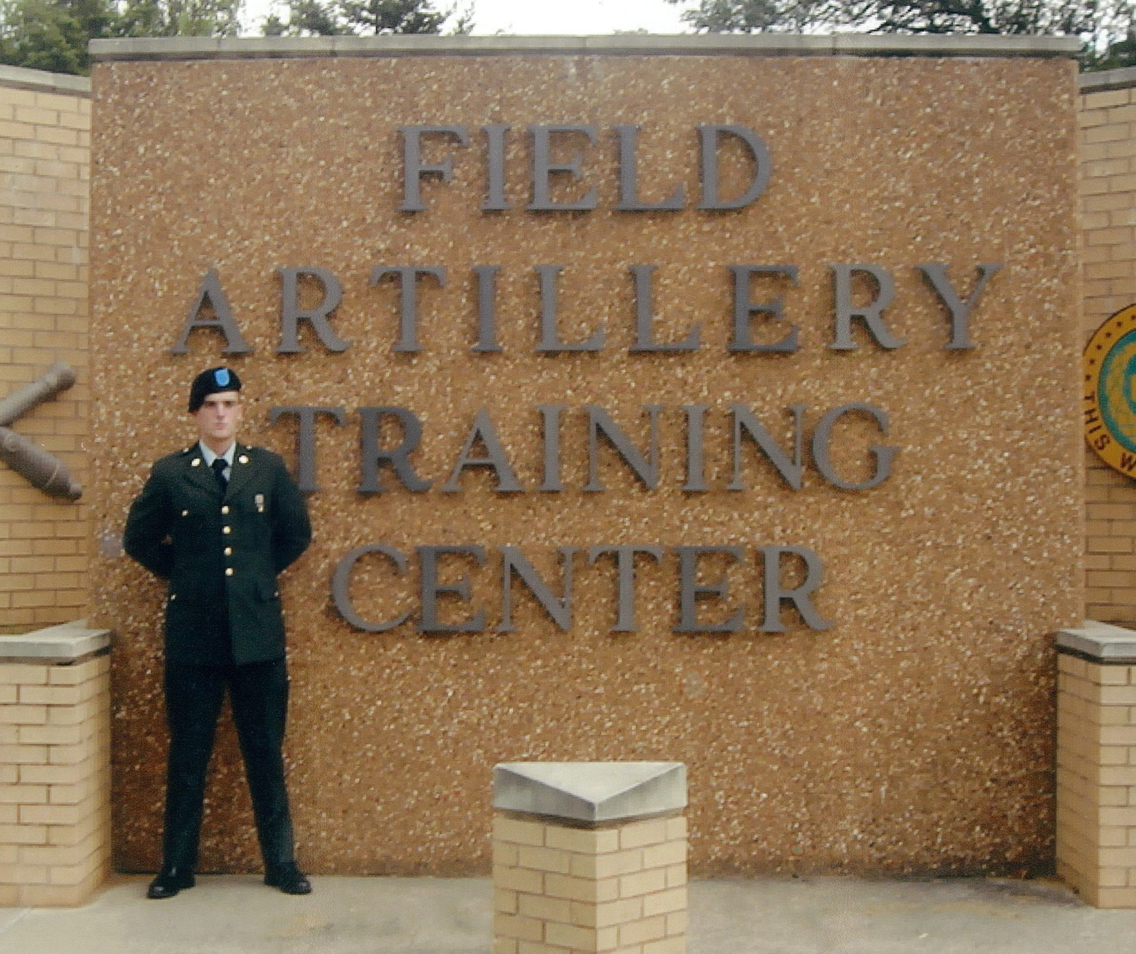 A uniformed soldier stands in front of a large sign that reads &quot;Field Artillery Training Center,&quot; made of textured brown material and flanked by brick pillars.