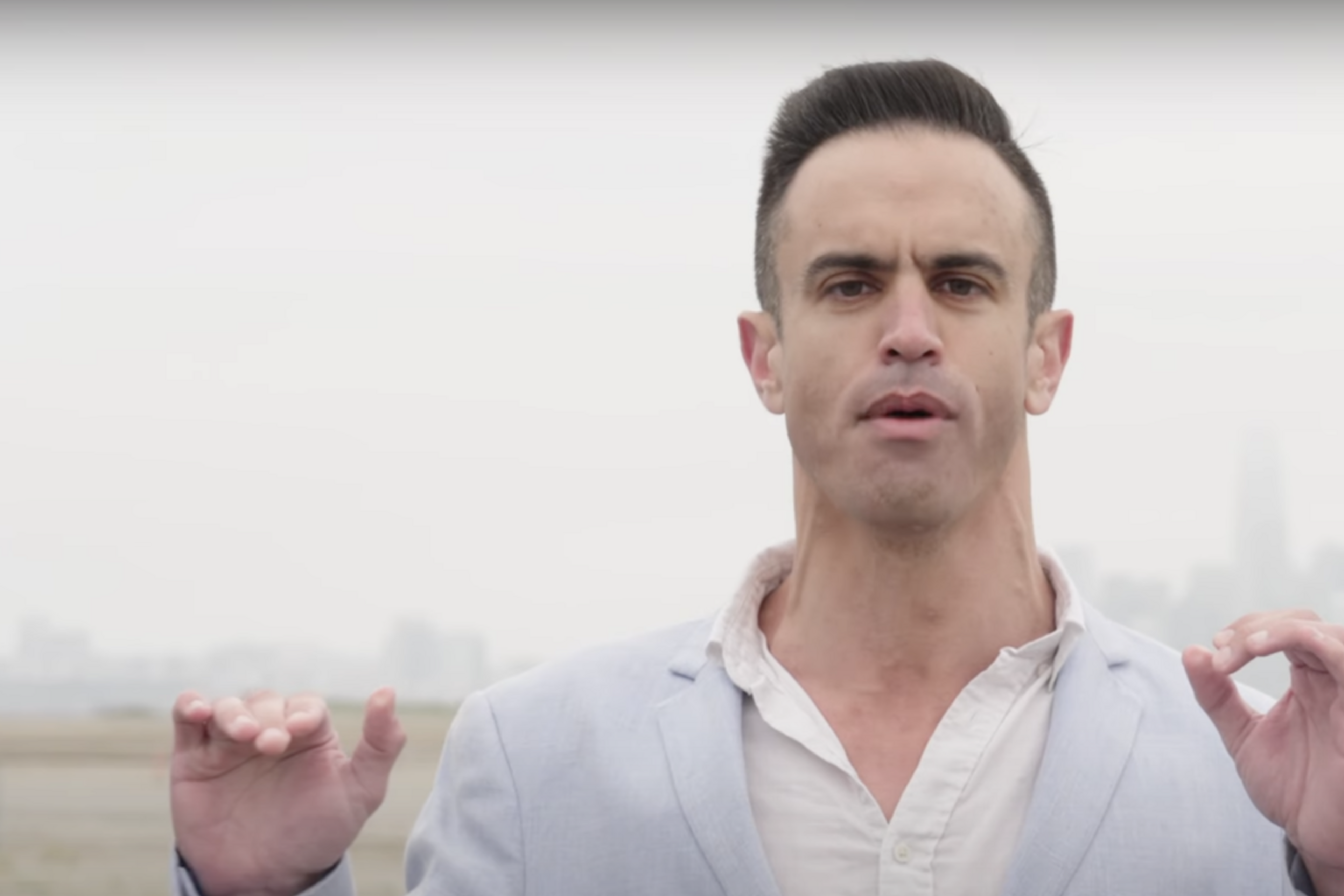 A person in a light blue suit stands outdoors with a city skyline in the background. Their hands are raised, and the text reads: "irredeemable aspects of modern society."