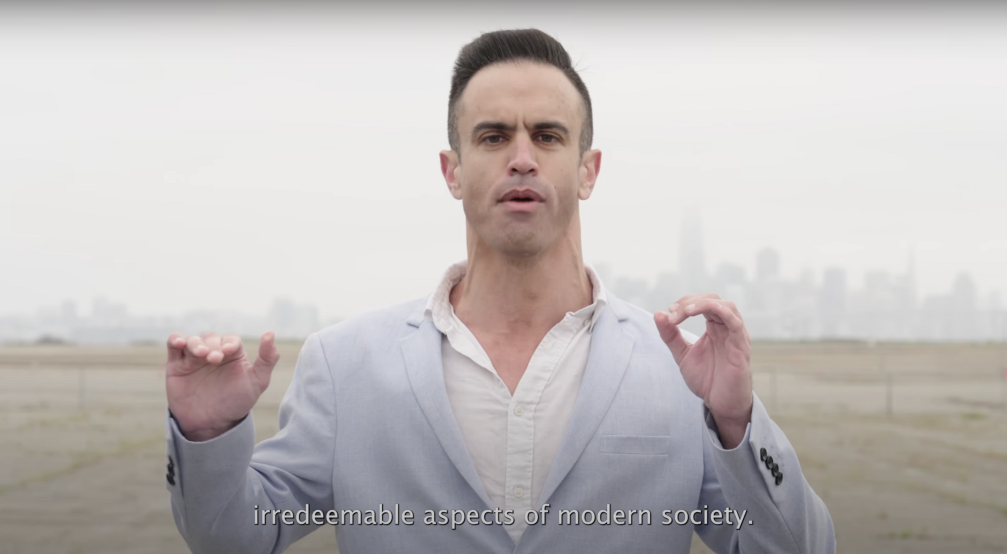 A person in a light blue suit stands outdoors with a city skyline in the background. Their hands are raised, and the text reads: "irredeemable aspects of modern society."