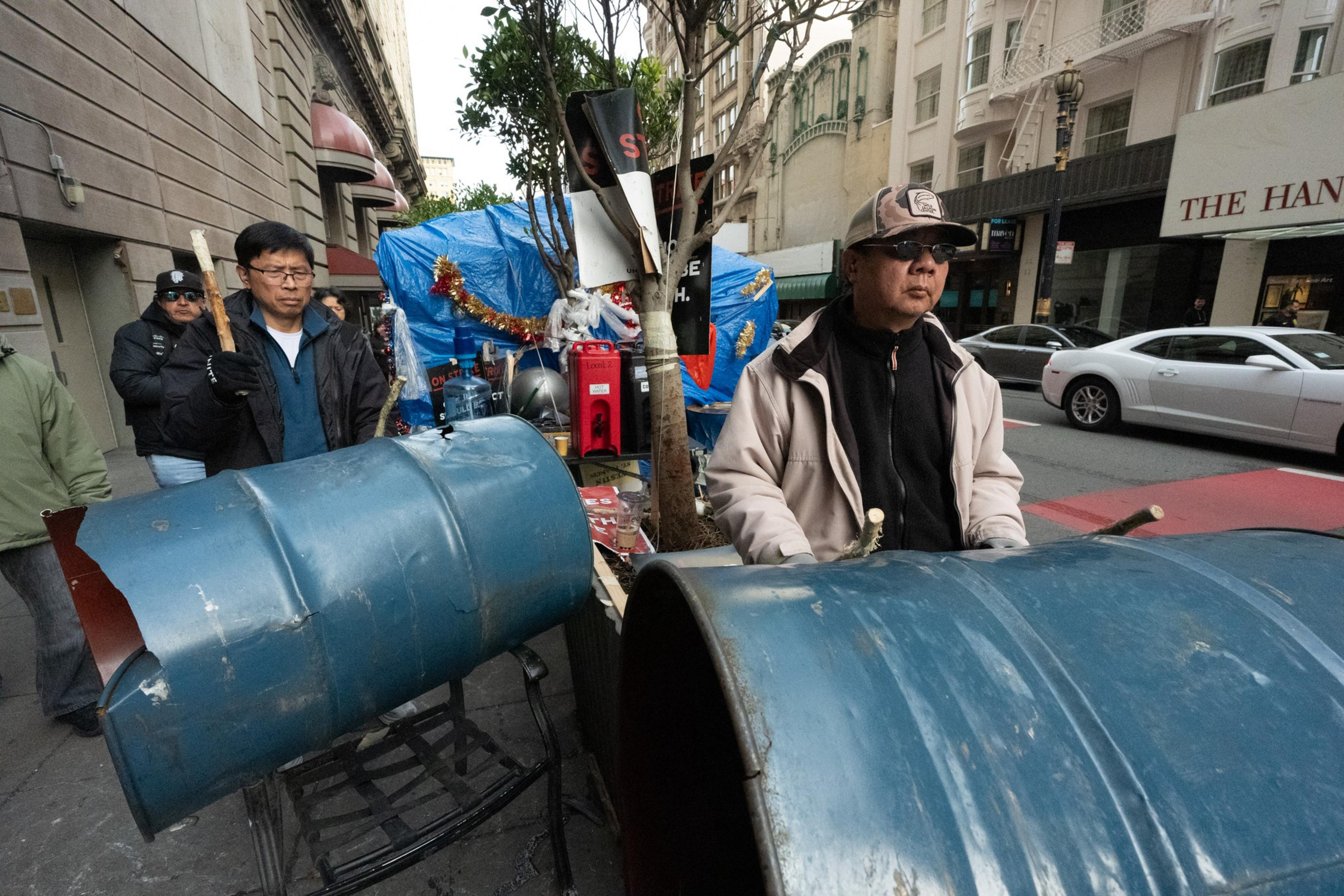 Two people are drumming on metal barrels on a city sidewalk with nearby pedestrians. A car is parked on the street, and there are blue tarps and decorations.