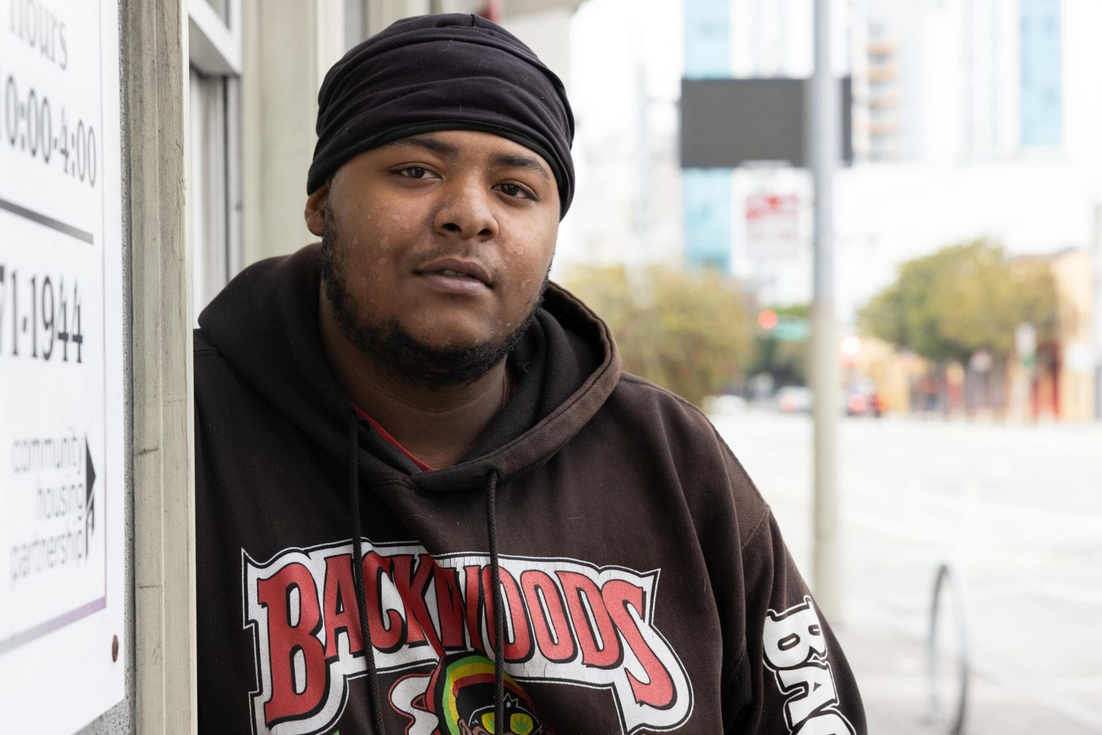A man wearing a black headwrap and a brown hoodie with "Backwoods" written on it leans against a wall on a city street with blurred buildings in the background.