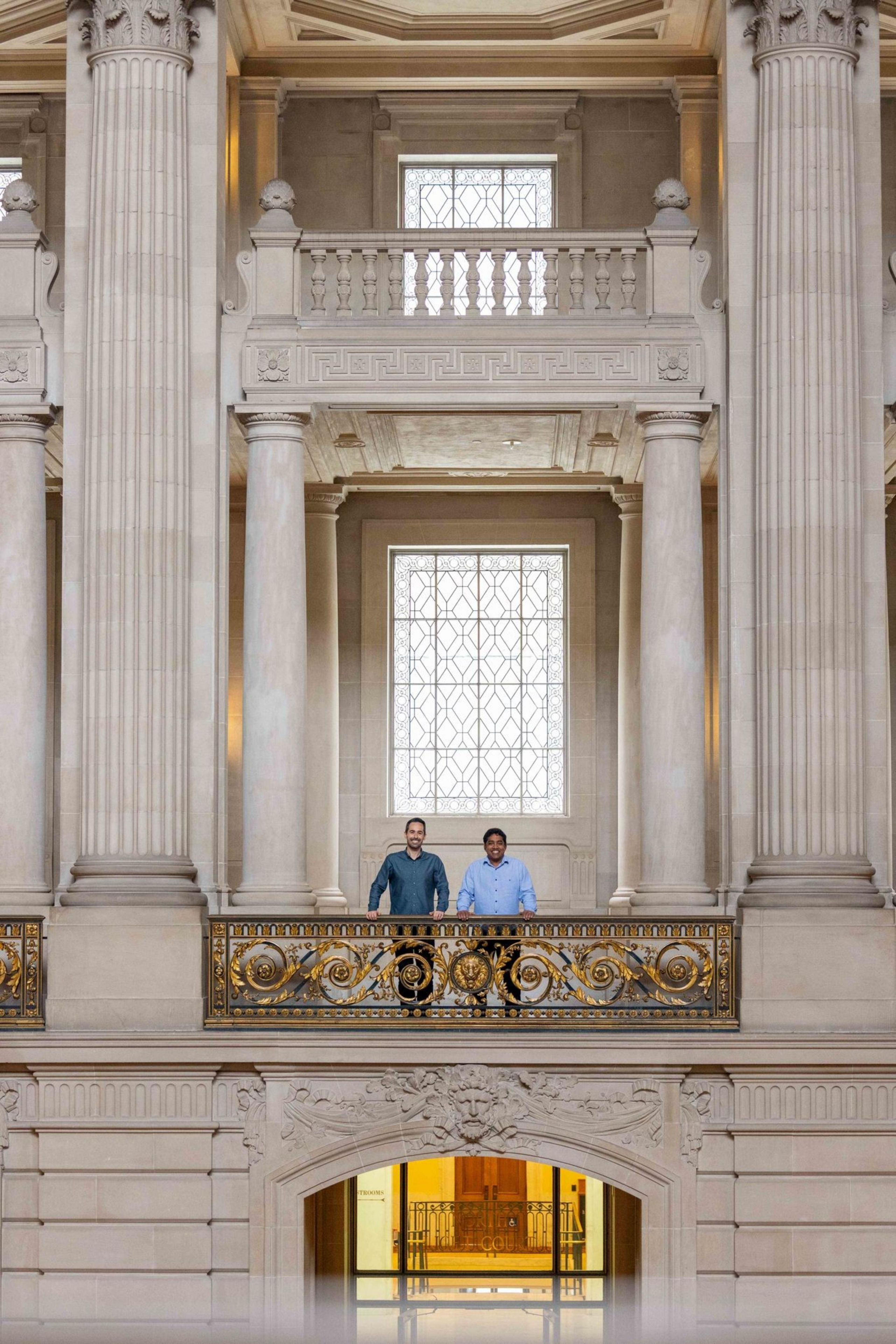 Two people stand on an ornate balcony in a grand building with large columns and decorative windows.