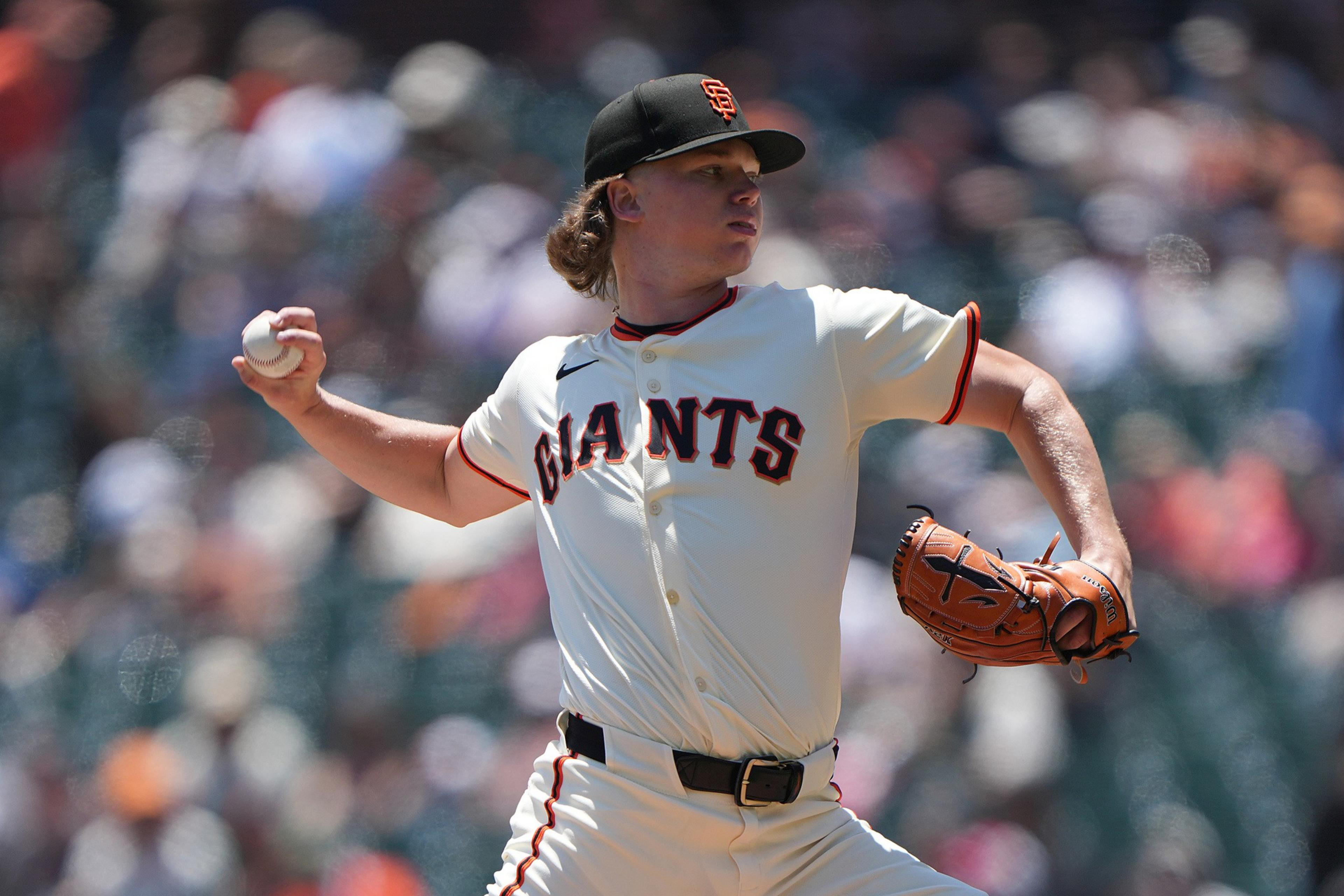 A baseball player in a "Giants" uniform is pitching on a sunny day, holding a ball in one hand and wearing a glove on the other, with a crowd blurred in the background.