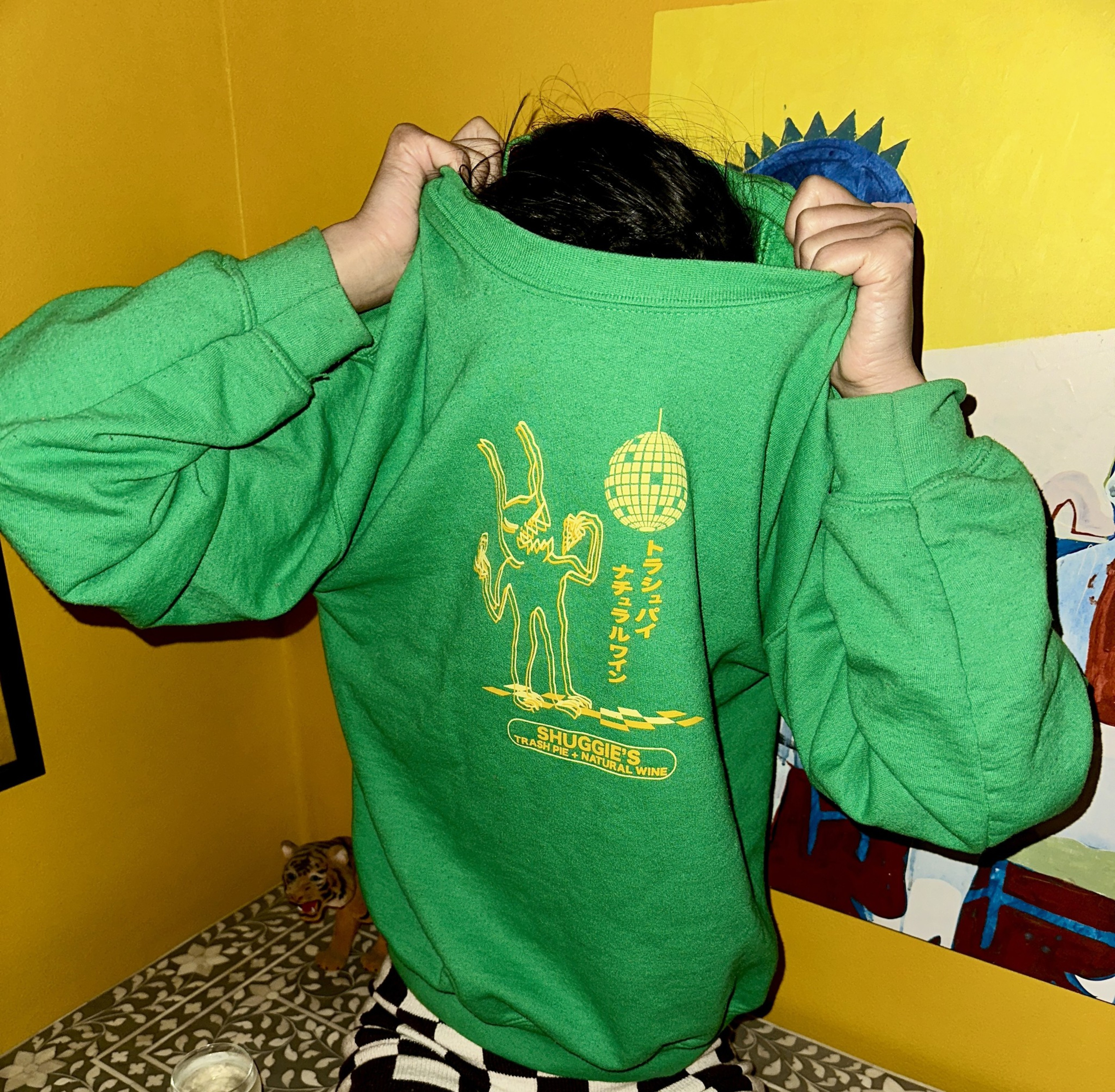 A person is pulling a green sweatshirt over their face. The shirt features a yellow graphic of a dancing figure, Japanese text, and the words &quot;Shuggie's Trash Pie &amp; Natural Wine.&quot;
