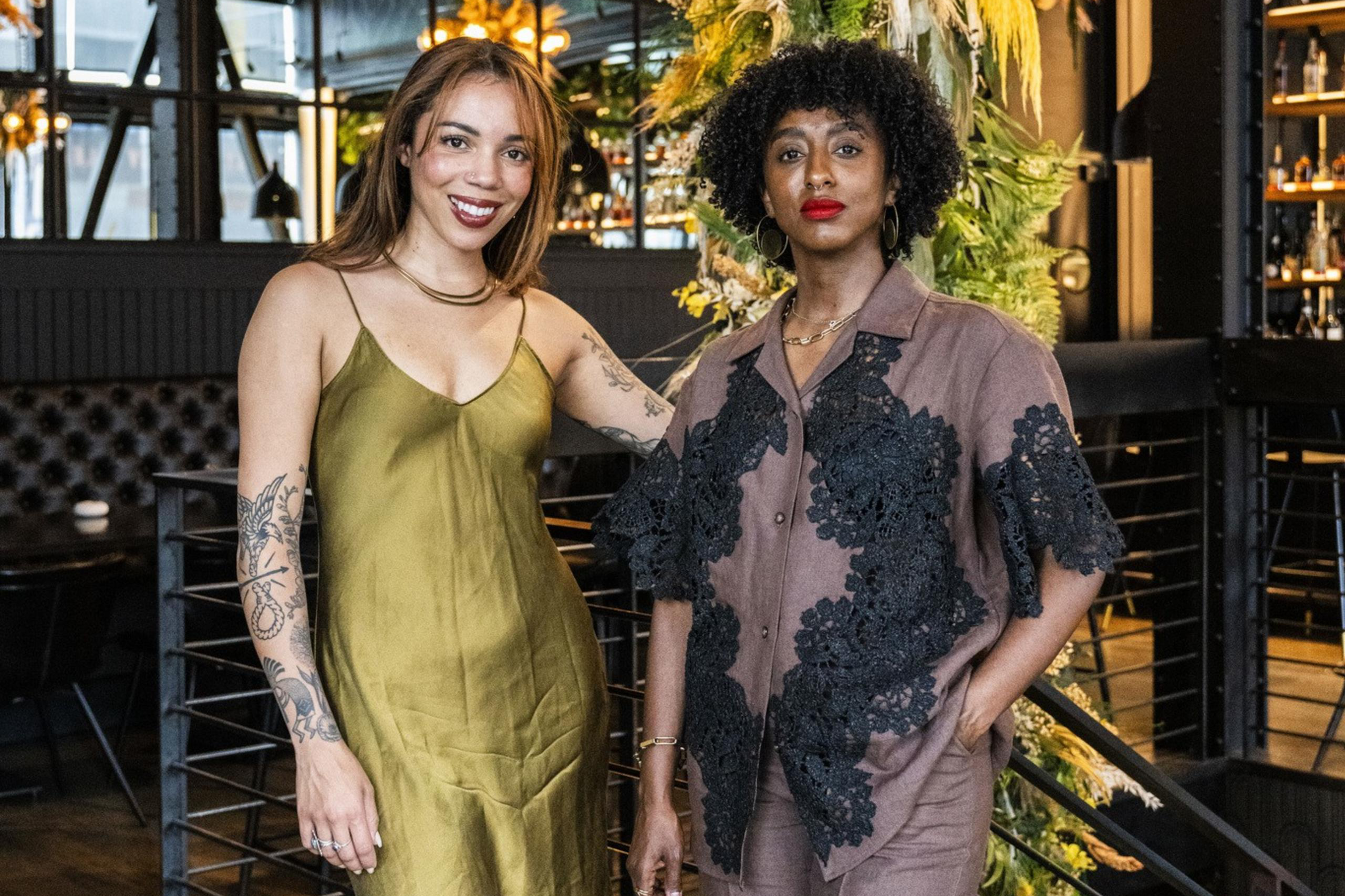 Two women stand indoors, one in a green dress, the other in a brown outfit with black patterns. They are beside a lush plant display and stylish lights.