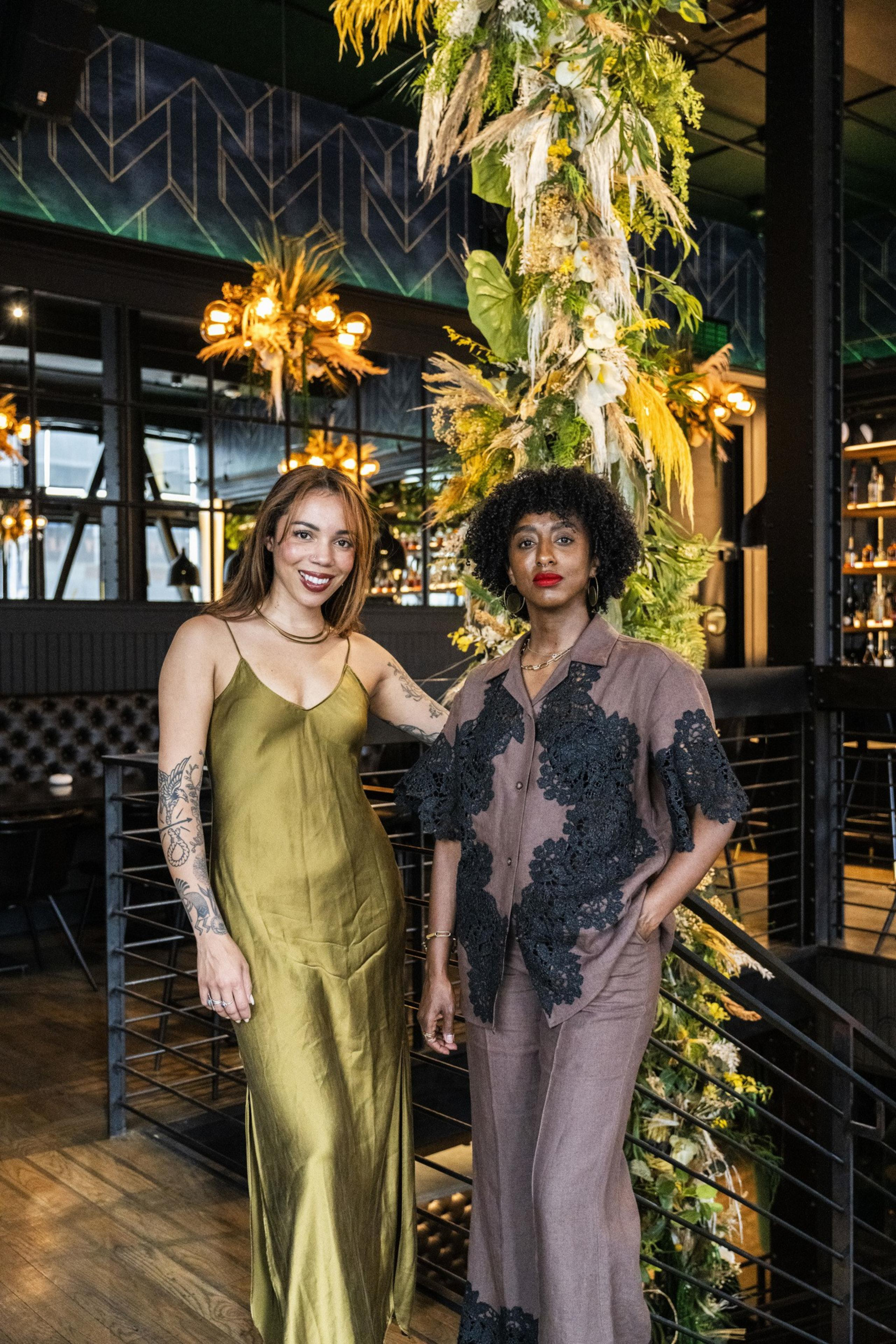 Two women stand indoors, one in a green dress, the other in a brown outfit with black patterns. They are beside a lush plant display and stylish lights.