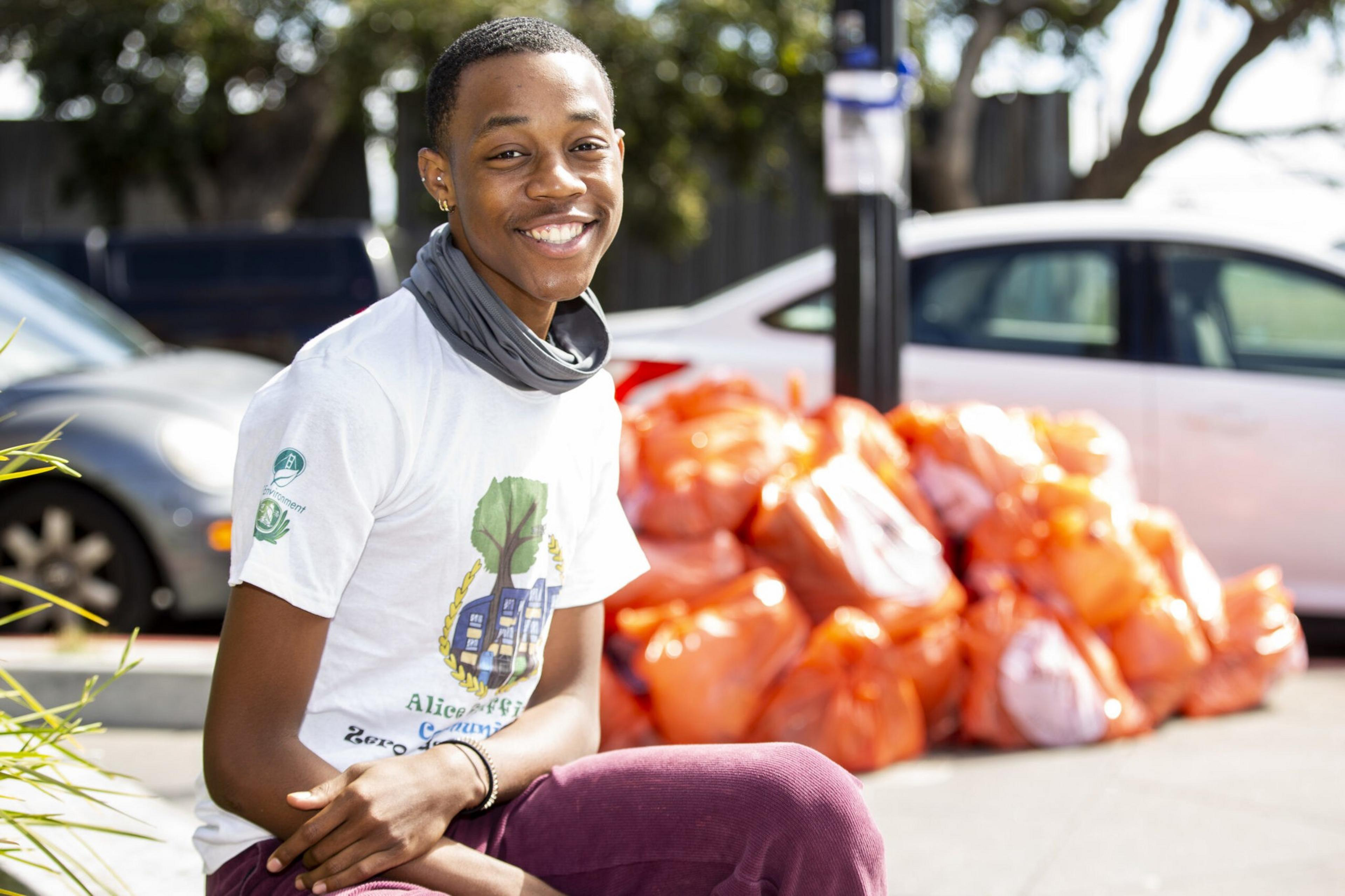 A young person smiles while sitting outdoors near a pile of orange garbage bags, with parked cars and trees in the background.
