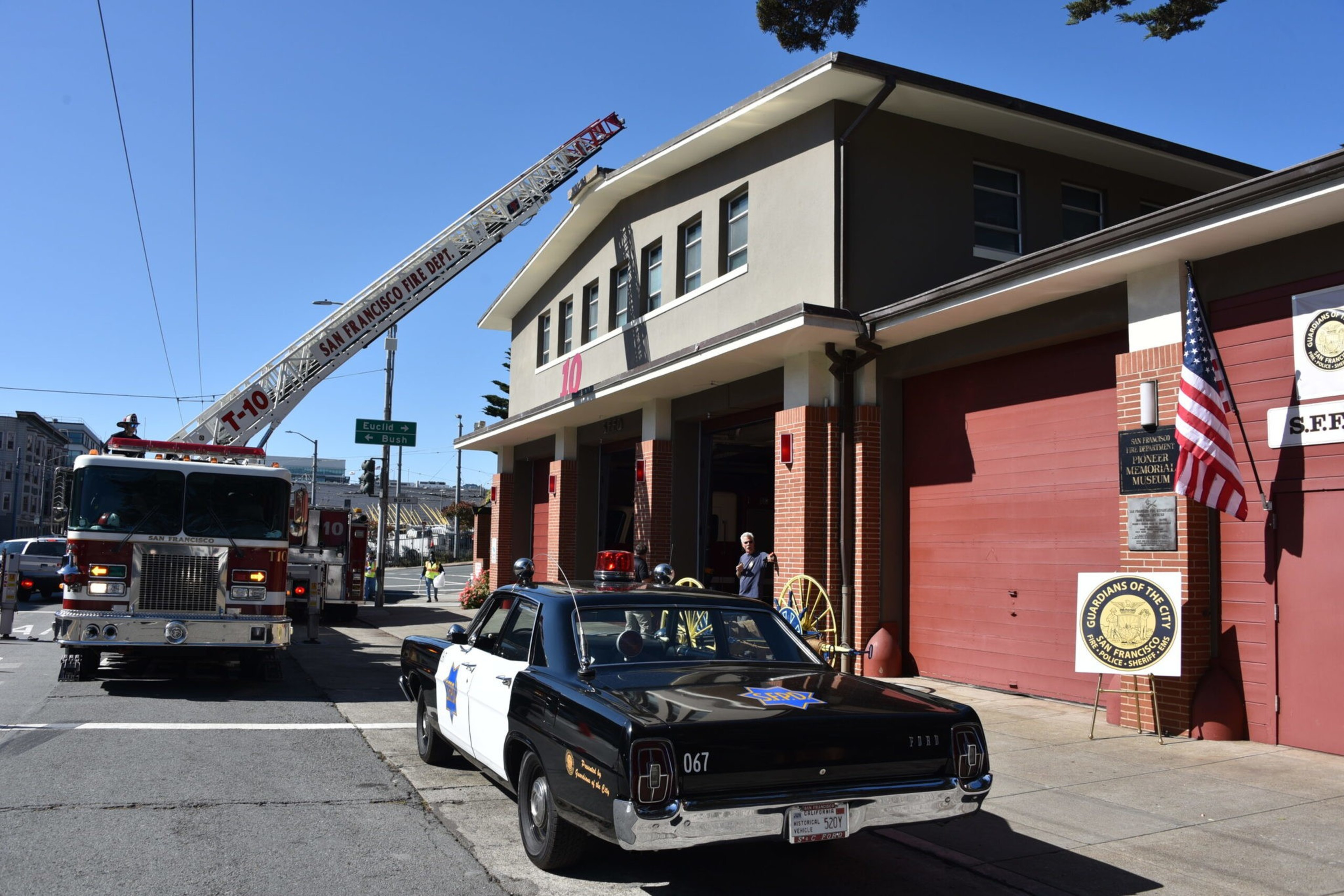 A vintage black-and-white police car is parked outside a fire station with a fire truck and ladder extended, an American flag, and signage visible.