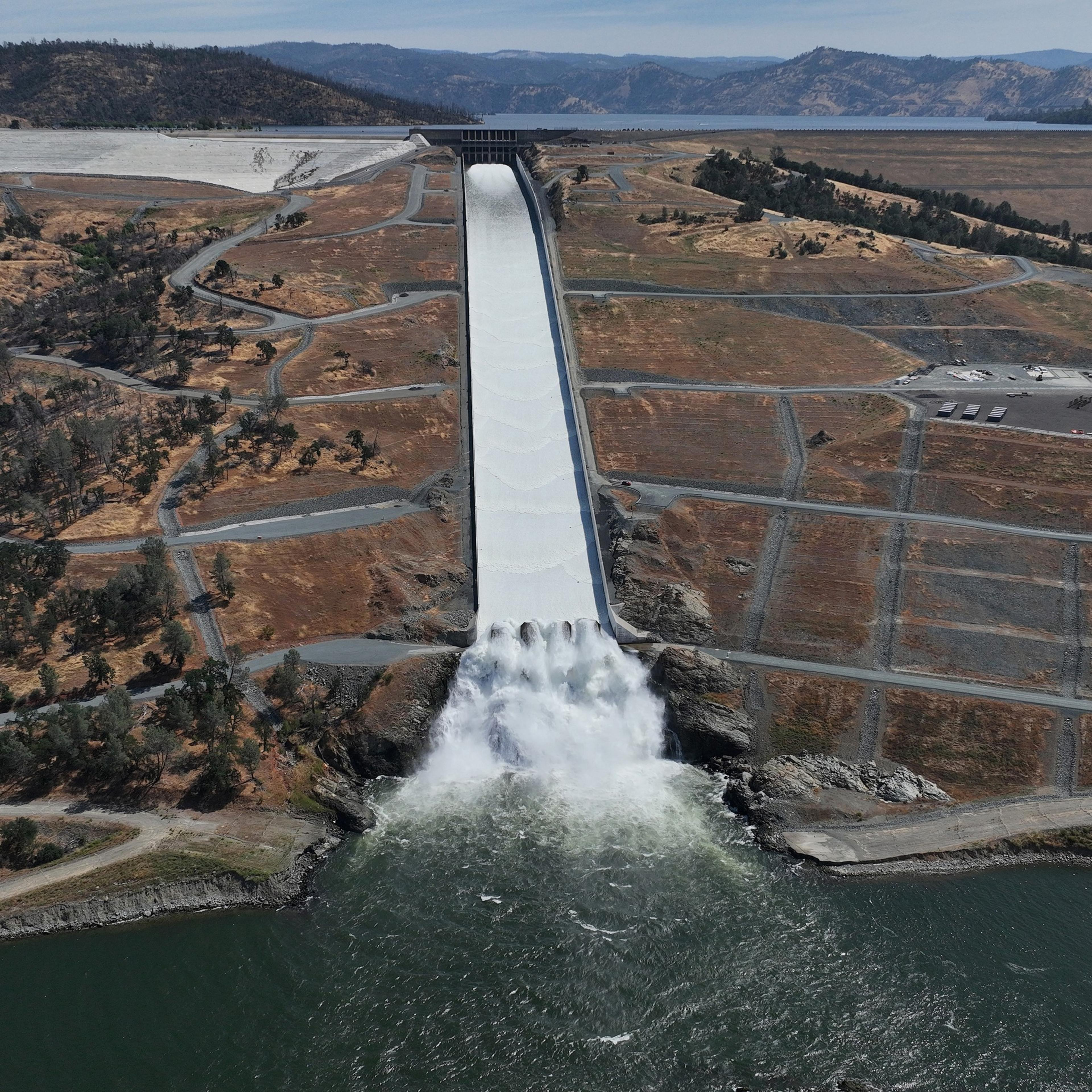 The image shows a dam with a massive spillway releasing water into a river. Surrounding the spillway are brown, dry lands with some scattered trees and roads.