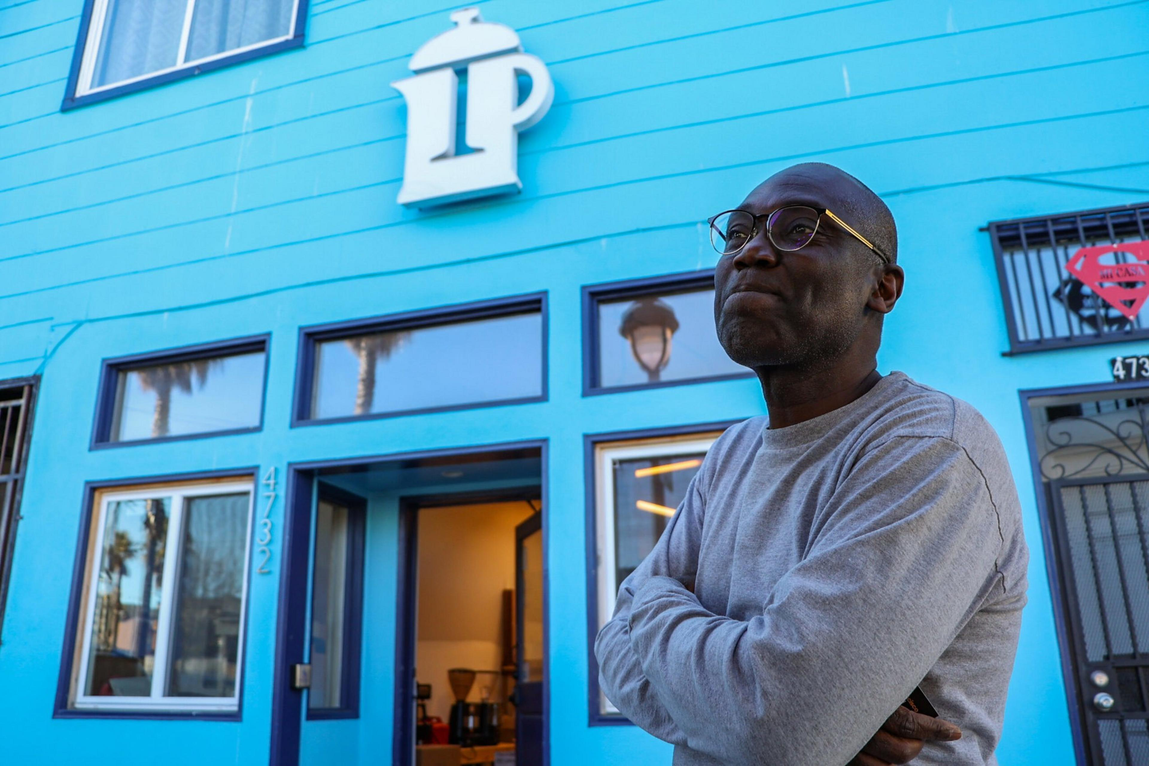 A man in glasses and a gray sweater stands with arms crossed outside a bright blue building with a coffee pot logo above the entrance.