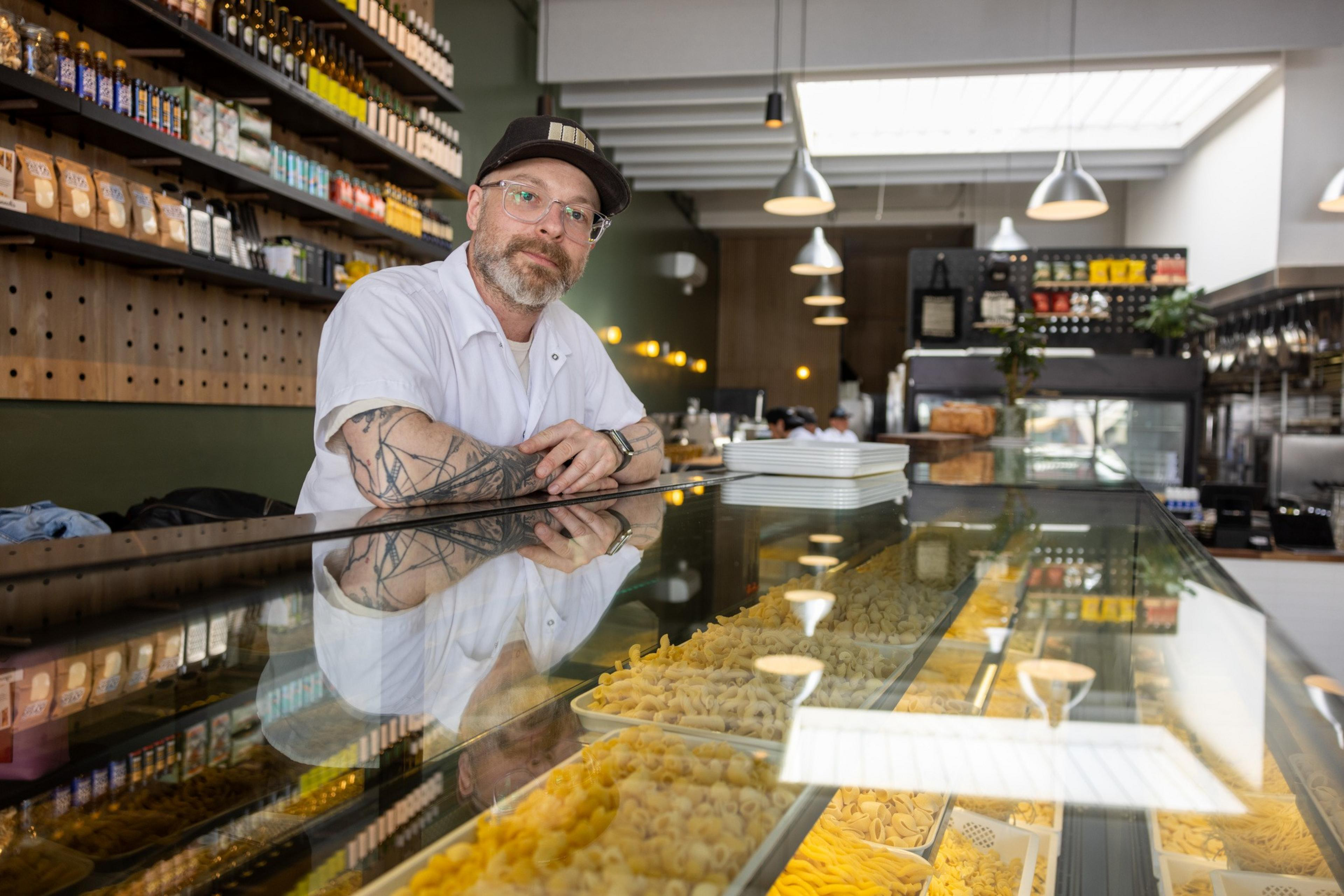 A bearded man with glasses and tattoos stands behind a glass counter displaying various types of pasta. Shelves with packaged goods and hanging lights are in the background.