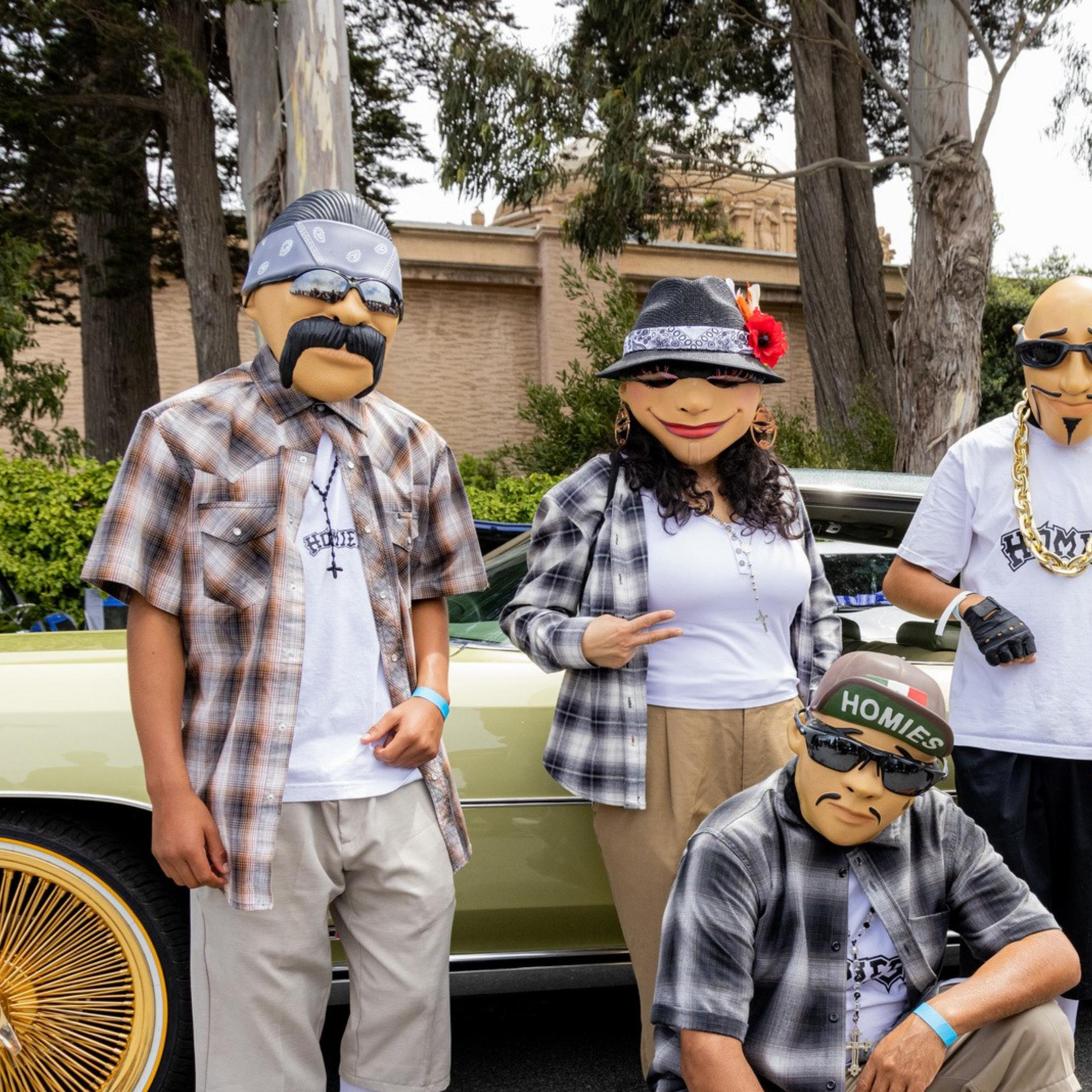 Five people wearing masks and casual clothing pose in front of a green vintage car with gold rims, outdoors on a street lined with trees, and spectators in the background.