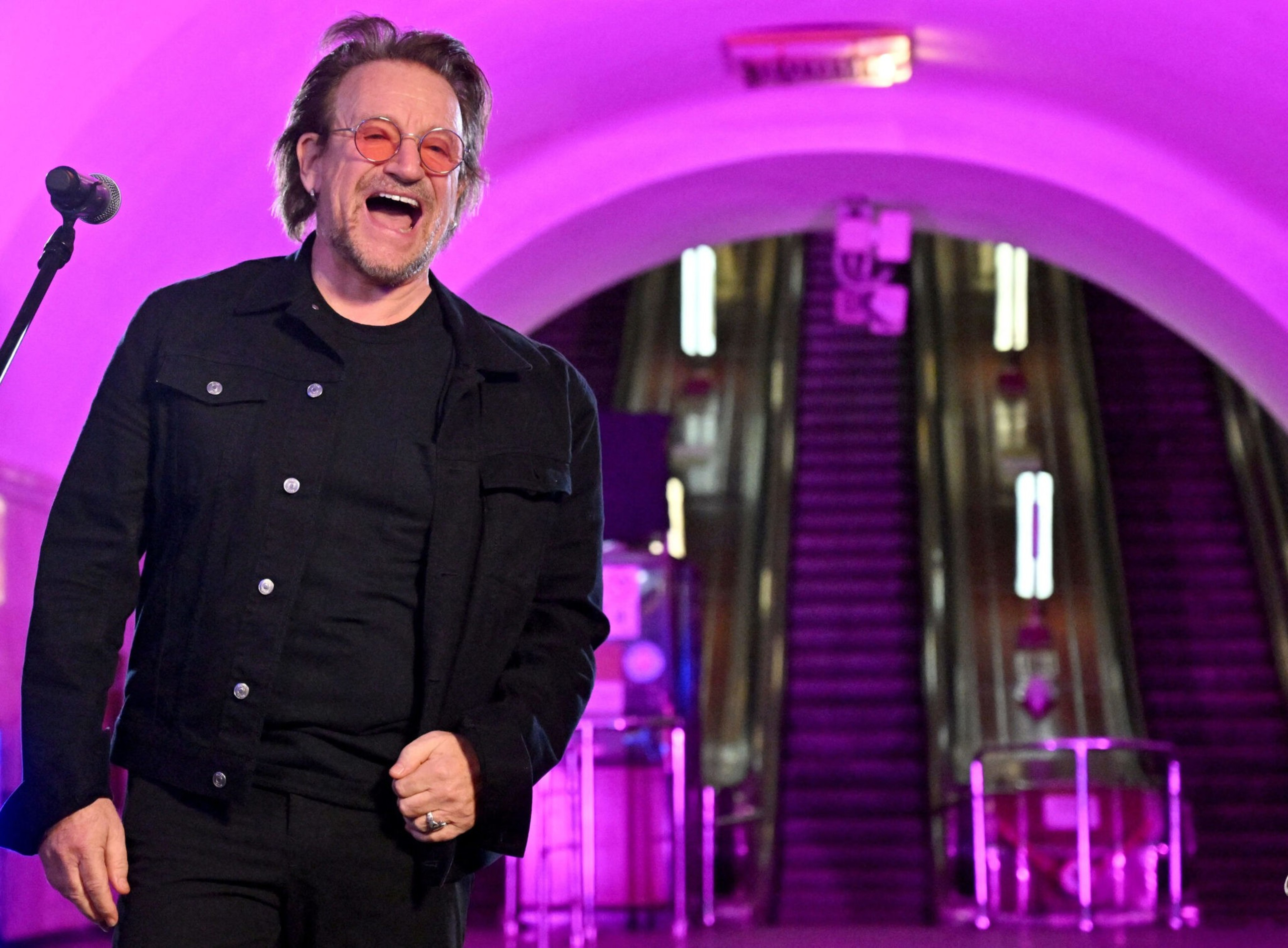 A man wearing pink-tinted glasses and a black jacket laughs near a microphone, with an escalator and purple lighting in the background.