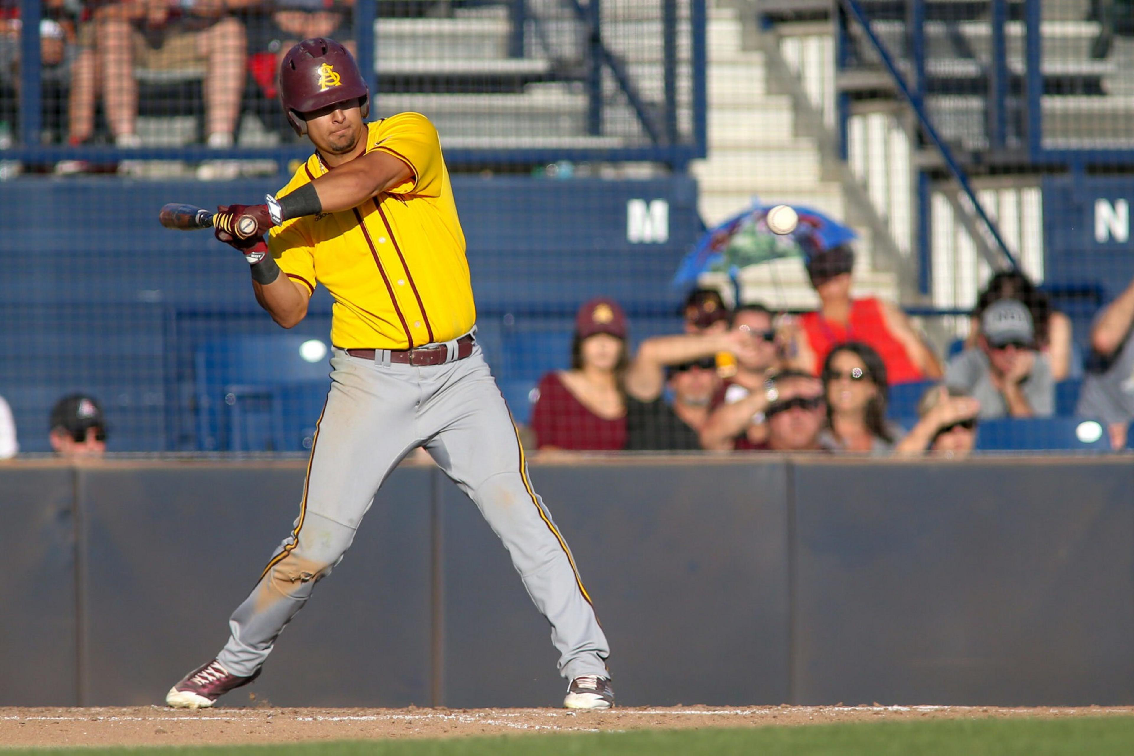 A baseball player in a yellow jersey and maroon helmet prepares to bunt as the ball approaches, with spectators watching behind a fence.