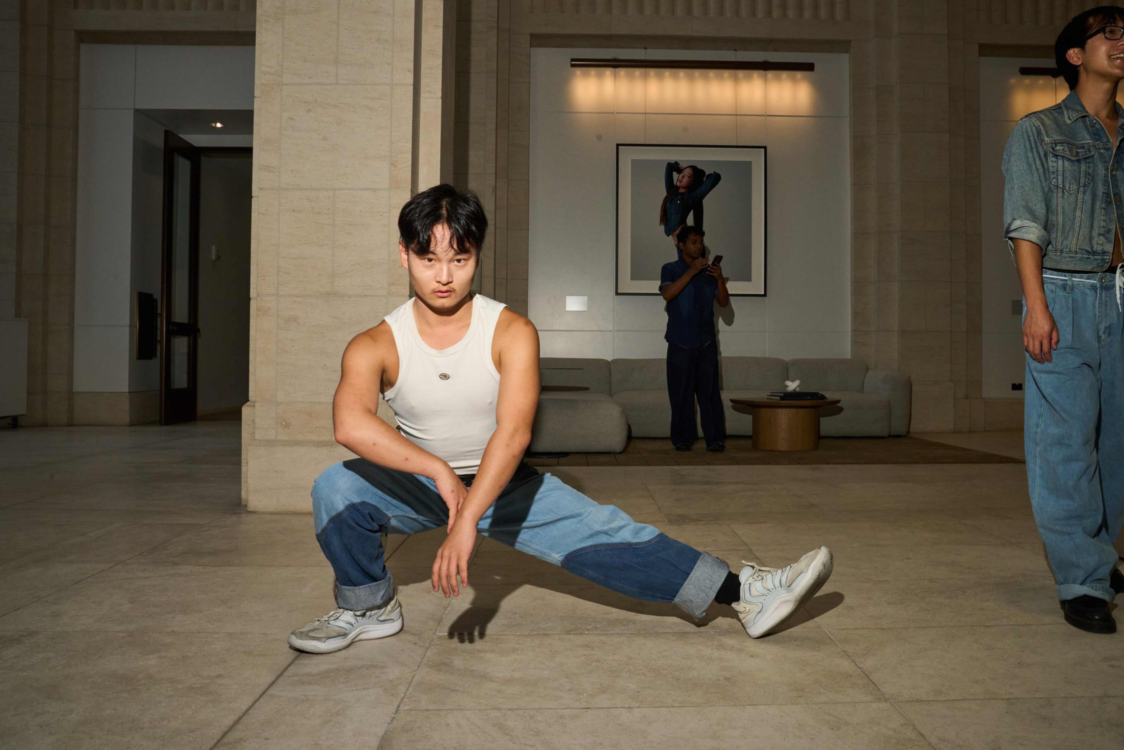 A young man in a white tank top and patchwork jeans stretches low with one leg bent and the other extended, while two others stand in the background.