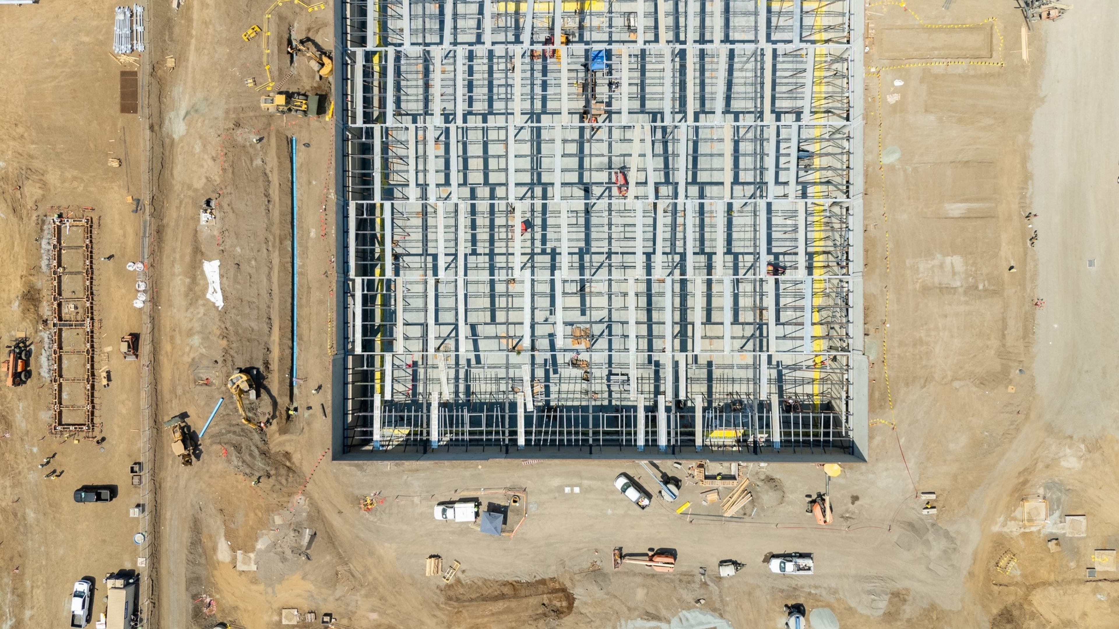 A large construction site with a rectangular steel framework building under construction, surrounded by dirt, construction vehicles, and equipment.