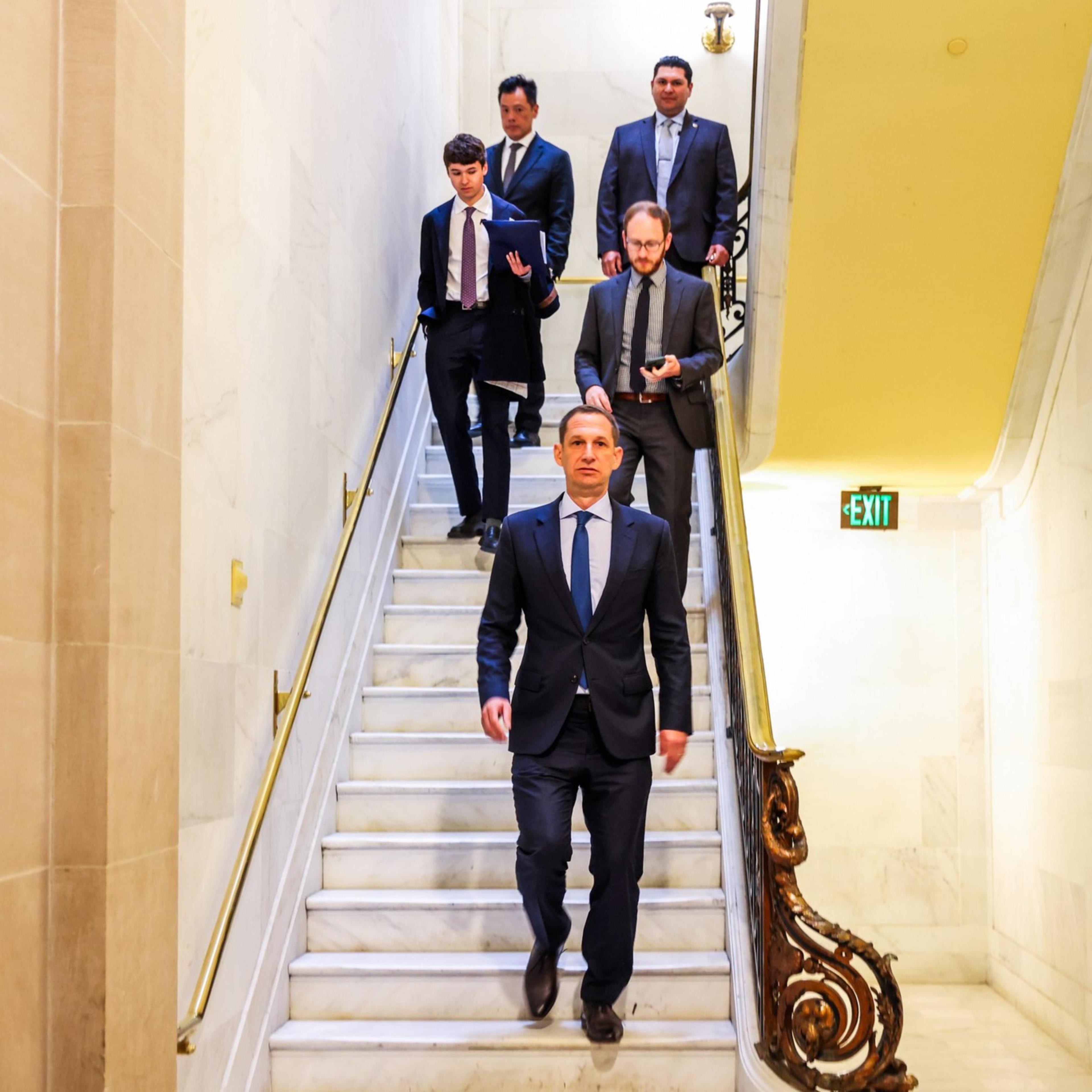 Five men in formal suits walk down a marble staircase with brass railings inside a bright, elegant building. An exit sign glows on the wall.