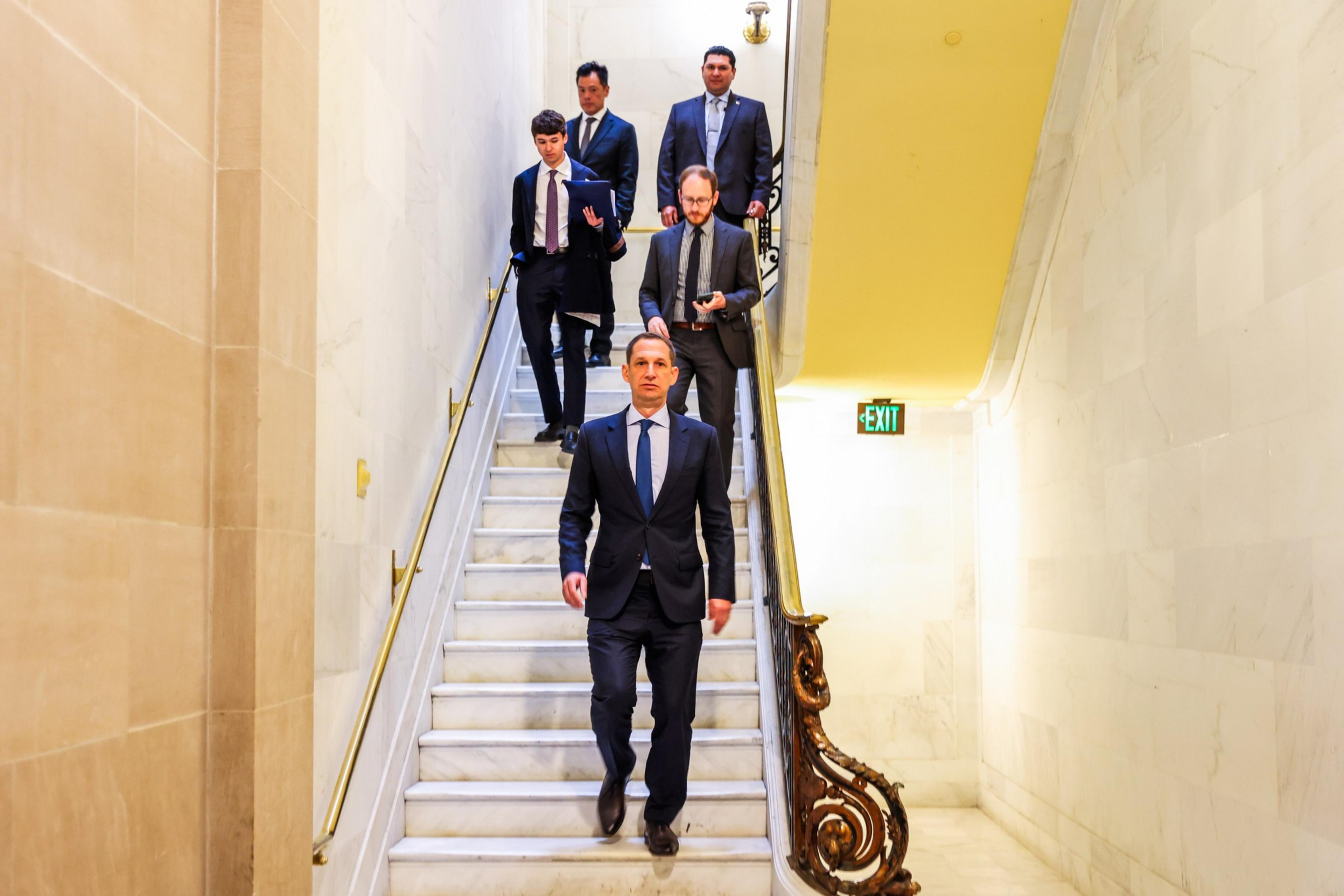 Five men in formal suits walk down a marble staircase with brass railings inside a bright, elegant building. An exit sign glows on the wall.